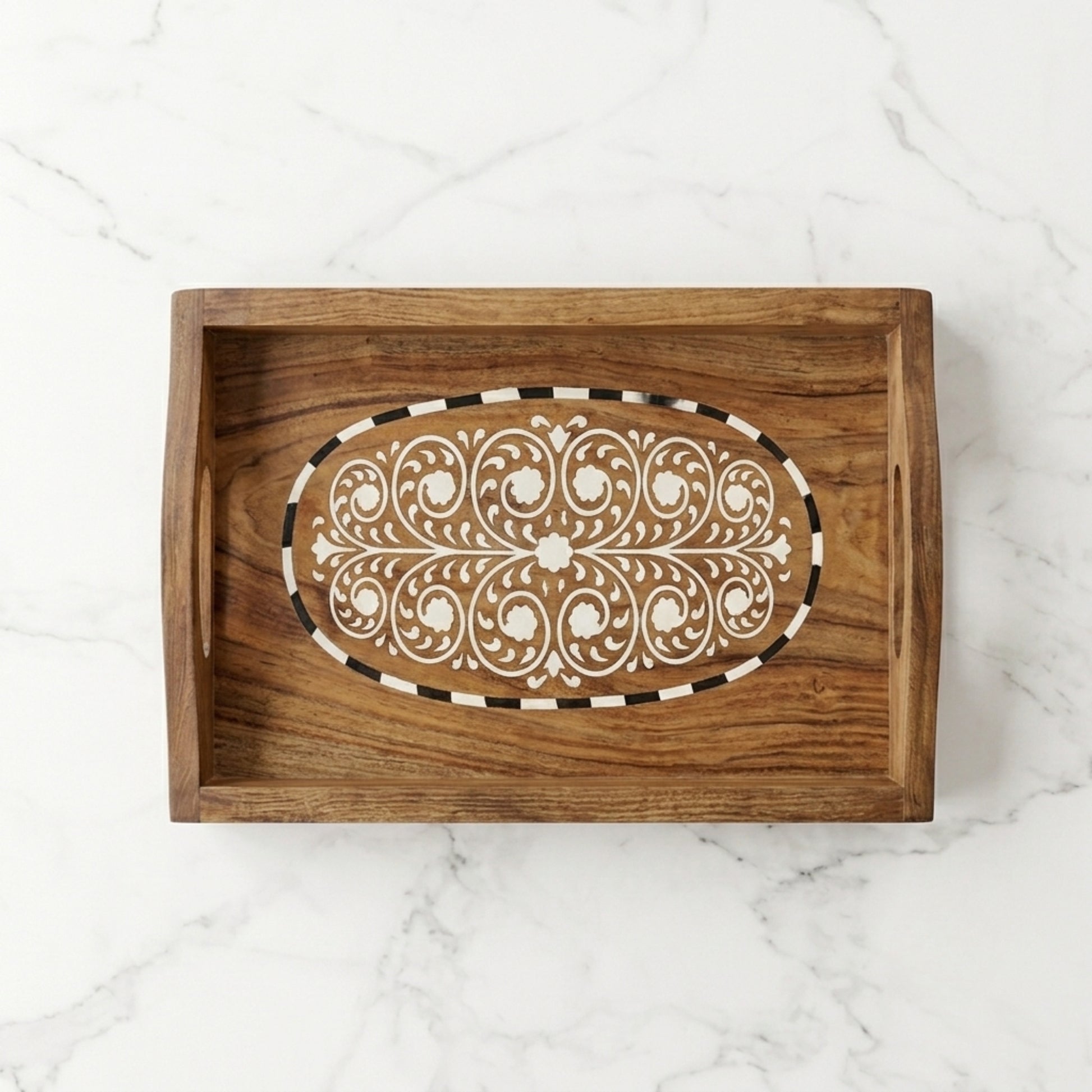 A rectangular wooden serving tray with a carved oval inlay featuring white floral scrollwork and a black-and-white striped border, shown on a white marble surface.