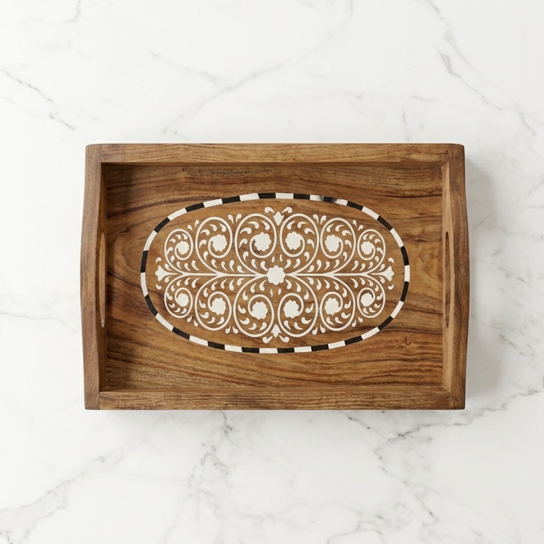 A rectangular wooden serving tray with a carved oval inlay featuring white floral scrollwork and a black-and-white striped border, shown on a white marble surface.