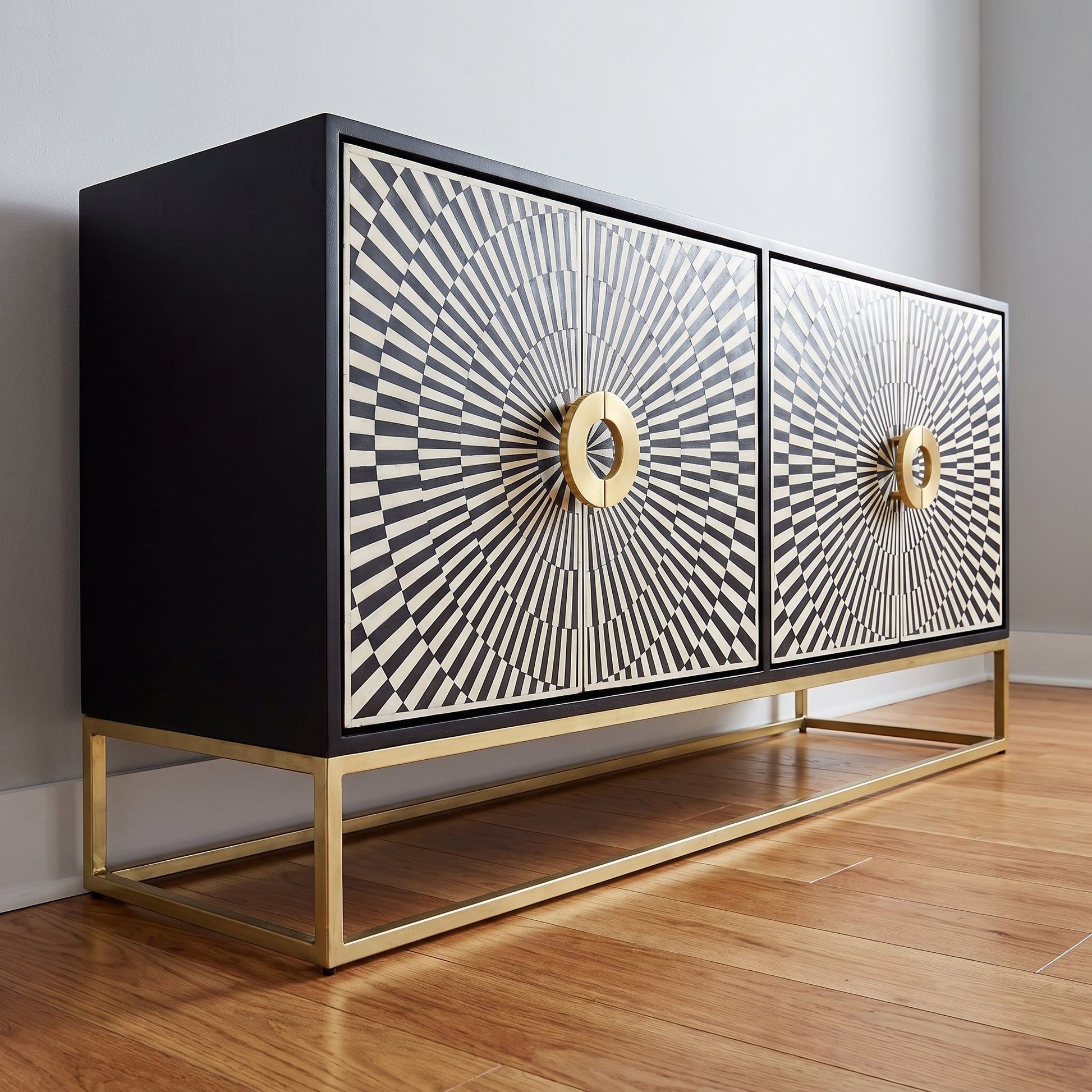 A modern two-door sideboard or credenza with a black frame, doors featuring a black-and-white sunburst pattern, and gold-tone circular handles, supported by a gold metal frame on a wood floor against a light gray wall.