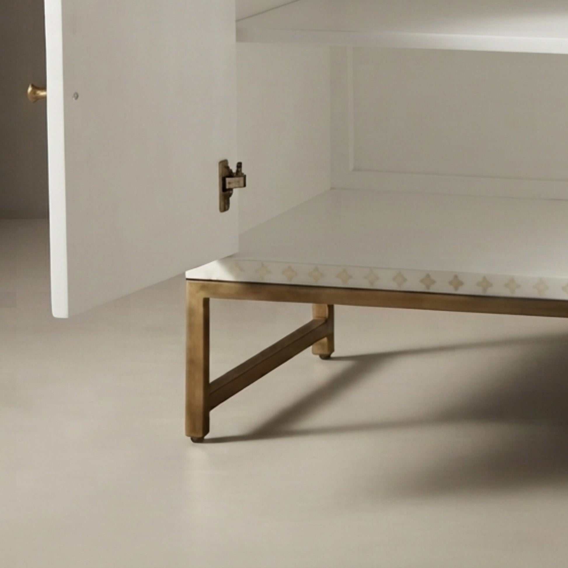 A close-up of a modern cabinet or shelf with a white interior, a white top surface, and a gold metal frame and legs. The cabinet door is slightly open, showing a brass hinge and knob.