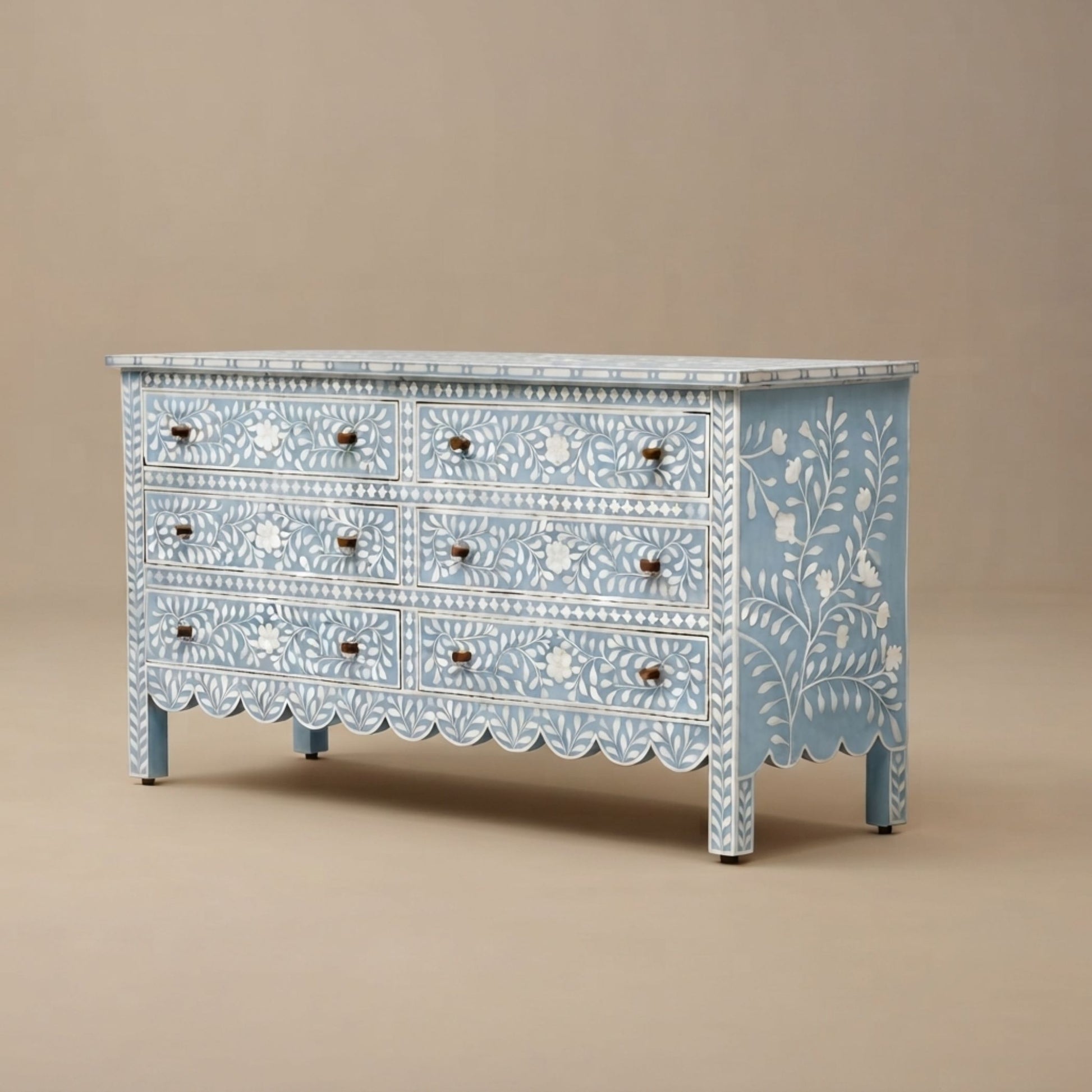 A light blue wooden dresser with white floral and geometric inlay patterns, featuring eight drawers with small round dark knobs and a scalloped front trim, set against a solid beige background.