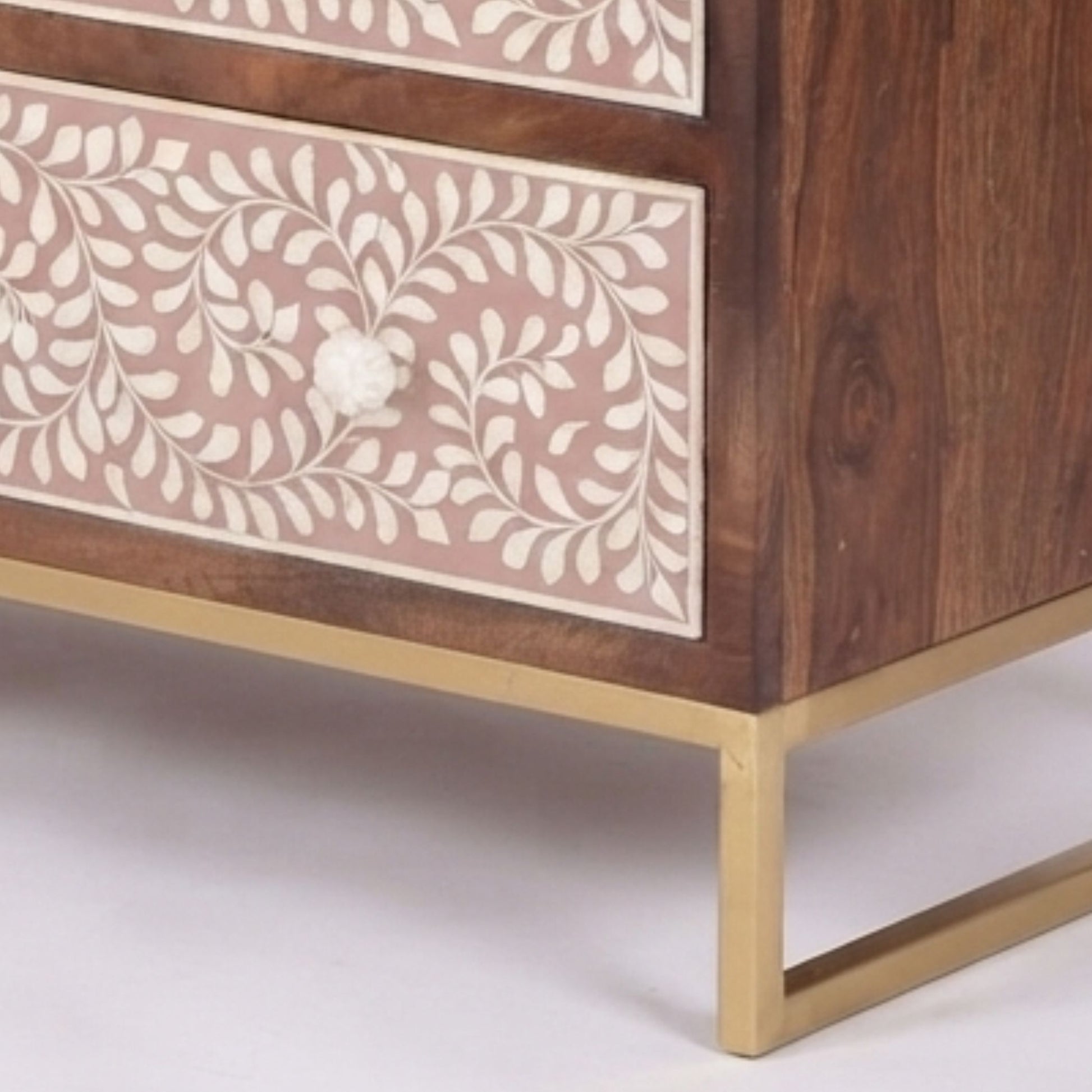 Close-up of a wooden chest of drawers with two drawers featuring cream bone-inlay floral patterns on a dusty pink background, accented with a white floral knob and a gold metal frame.