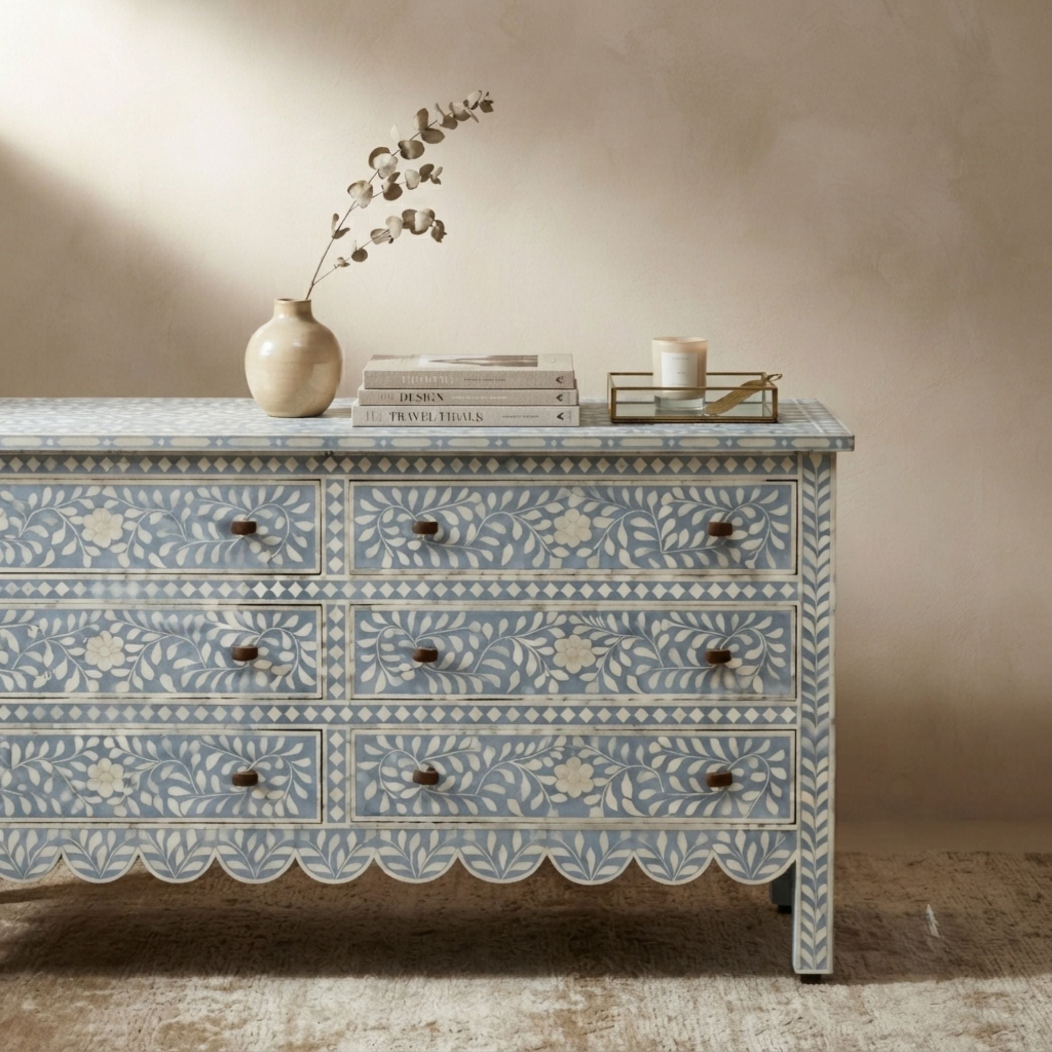 A light blue and white bone-inlay dresser with scalloped drawer fronts, styled with a ceramic vase, books, and a candle on top against a textured beige wall.