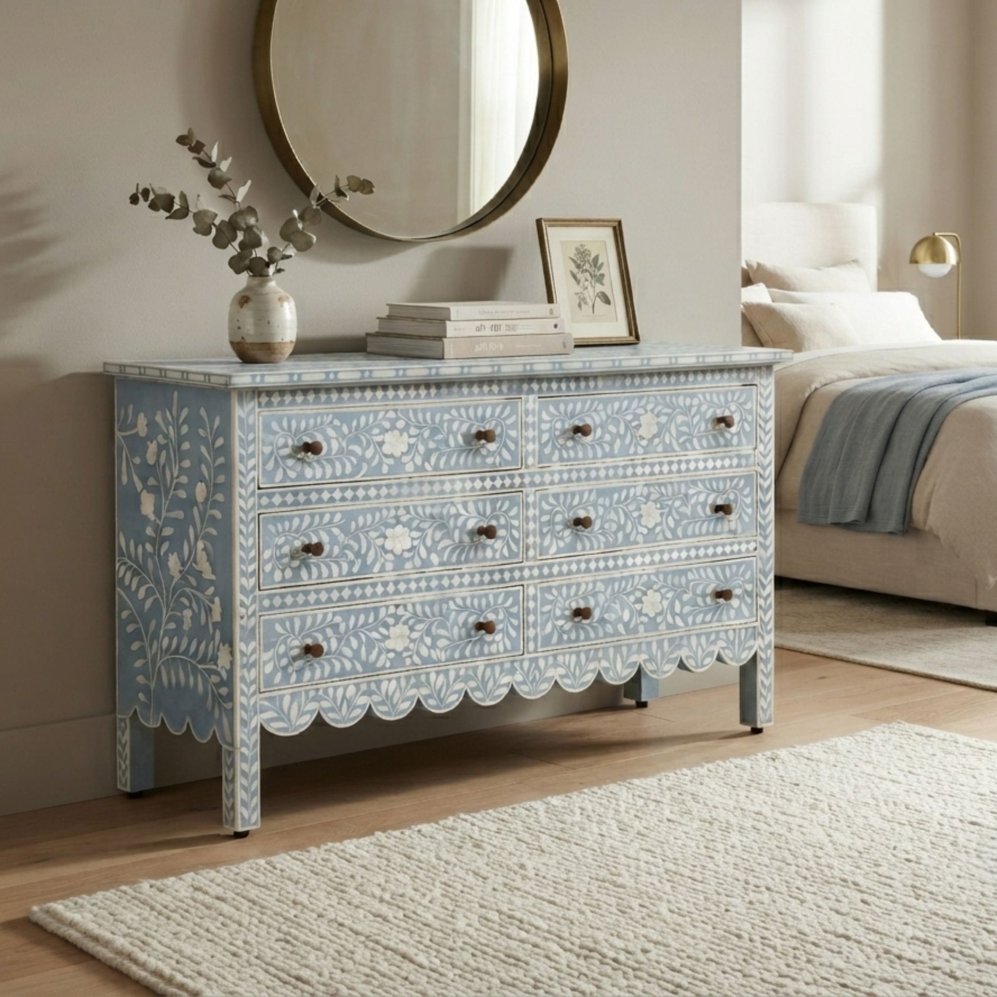A light blue and white patterned wooden dresser with scalloped drawer fronts and dark metal knobs, styled in a bedroom setting next to a bed and under a round mirror.