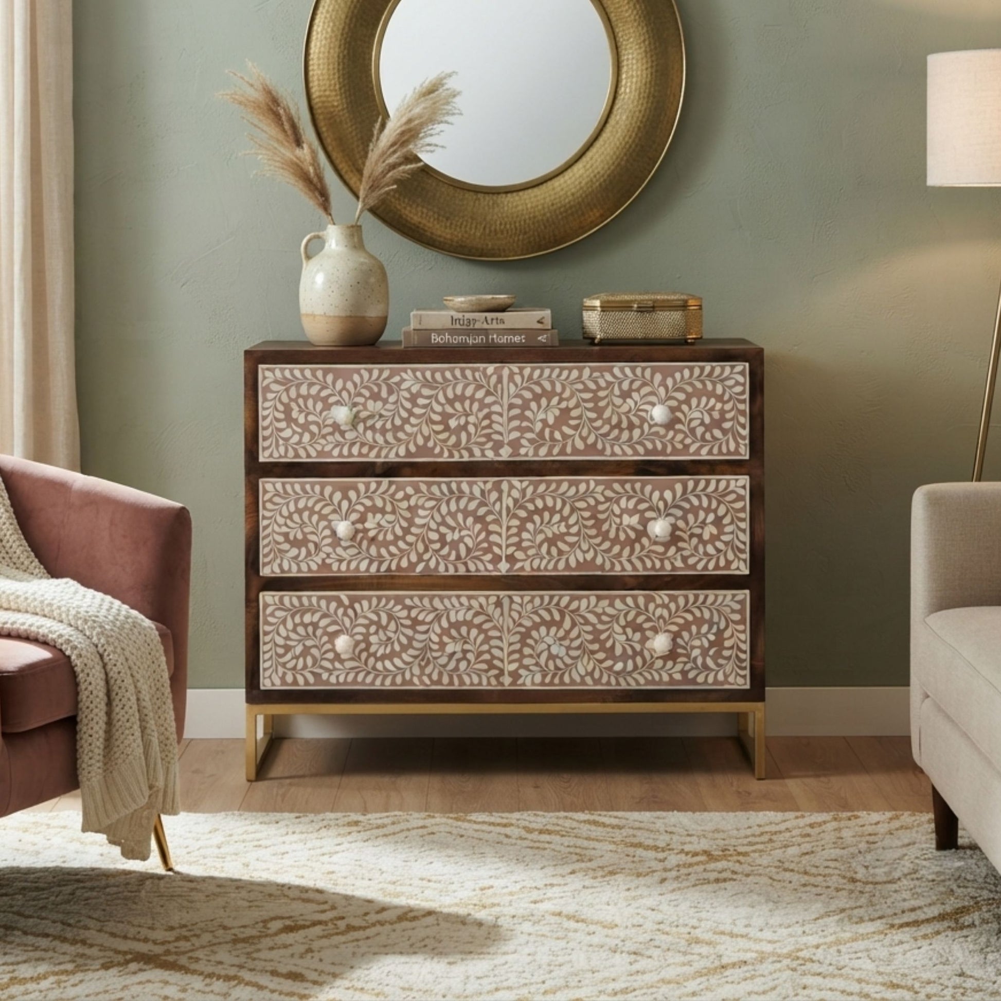 A three-drawer chest of drawers with carved floral-patterned beige drawers and dark brown wood trim, styled in a living room setting with a round gold mirror, vase, and books on top.