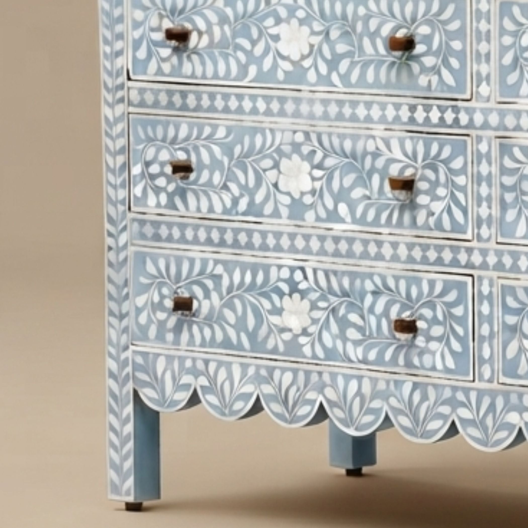 A close-up of a light blue and white carved wooden chest of drawers with floral patterns and scalloped drawer fronts, set against a neutral beige background.