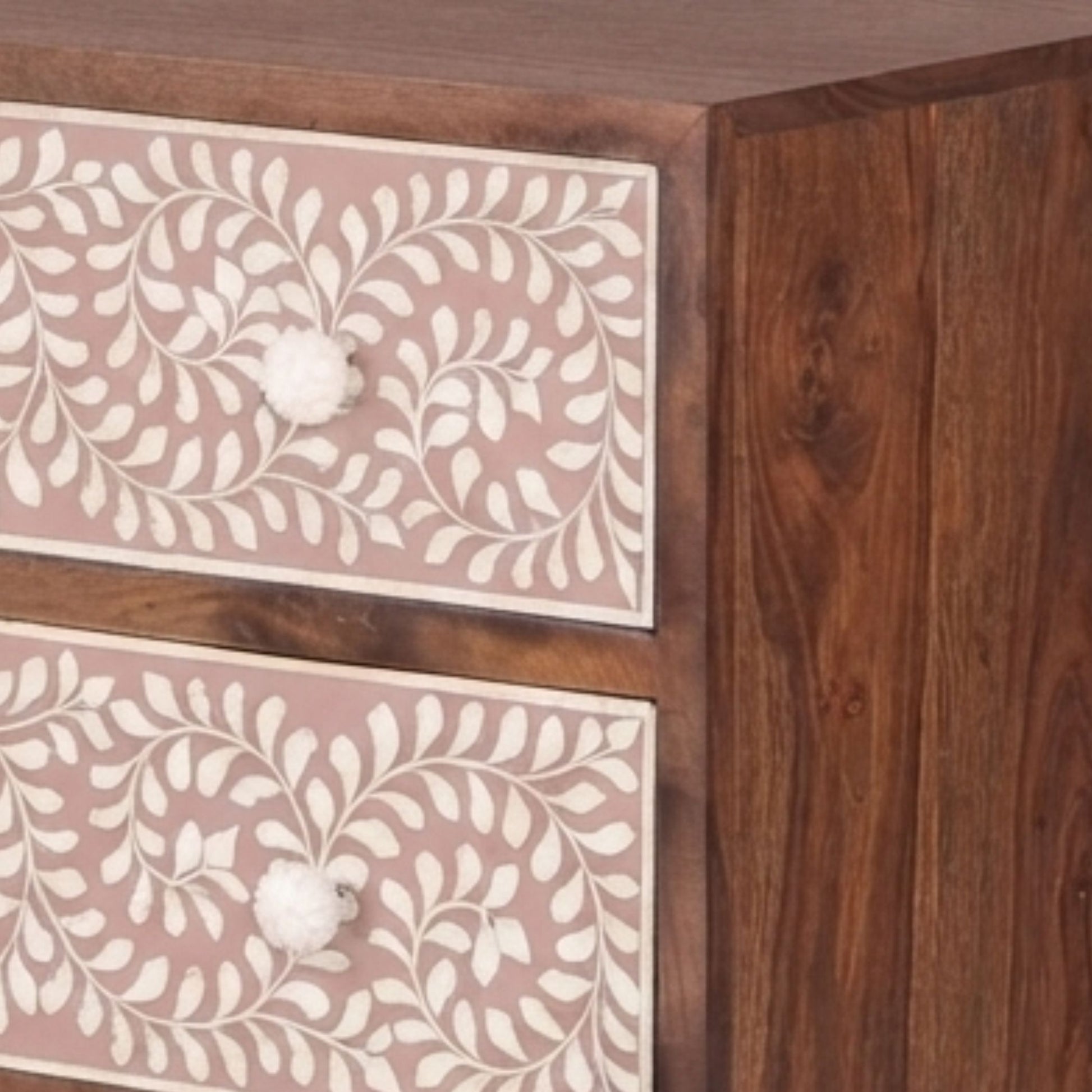 Close-up of a wooden cabinet or dresser with two drawers featuring dusty pink panels decorated with white floral vine patterns and round white knobs, set against a dark brown wood grain body.