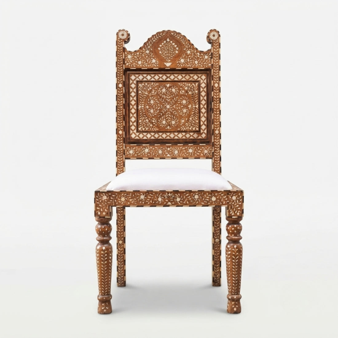 An ornate carved wooden chair with a white upholstered seat is centered in a richly decorated bohemian-style room with patterned rugs, cushions, lanterns, framed wall art, and plants.