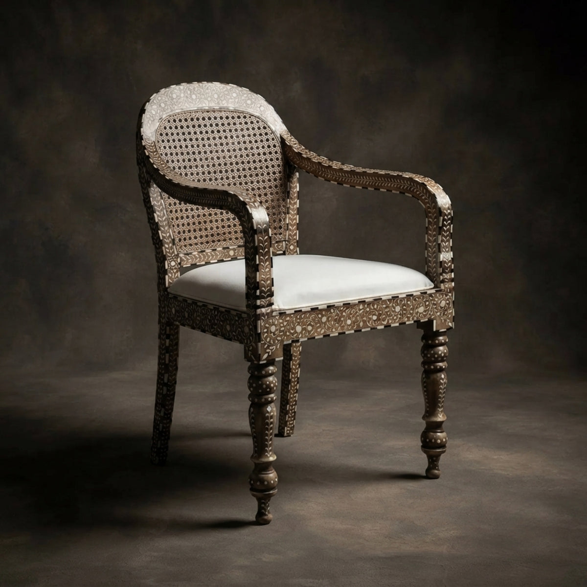 An ornate vintage-style armchair with a woven cane backrest, carved dark wood arms and legs, and a light beige upholstered seat cushion, photographed against a dark textured studio backdrop.