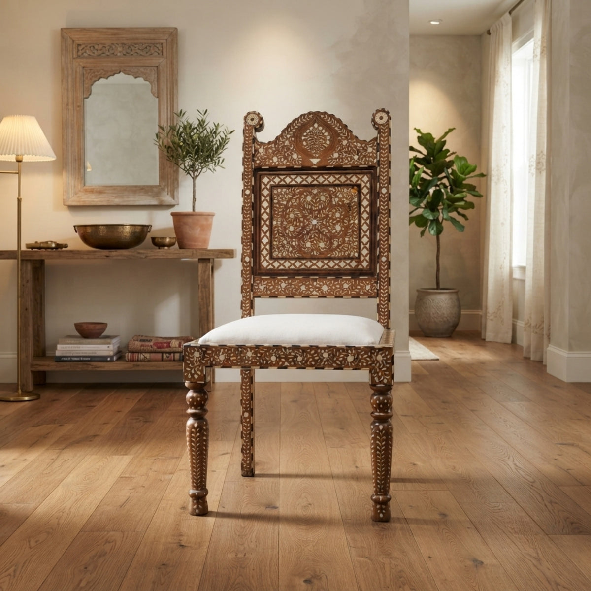 An ornate carved wooden chair with a white upholstered seat, placed in a warm, styled interior room with wood floors, a console table, and potted plants.