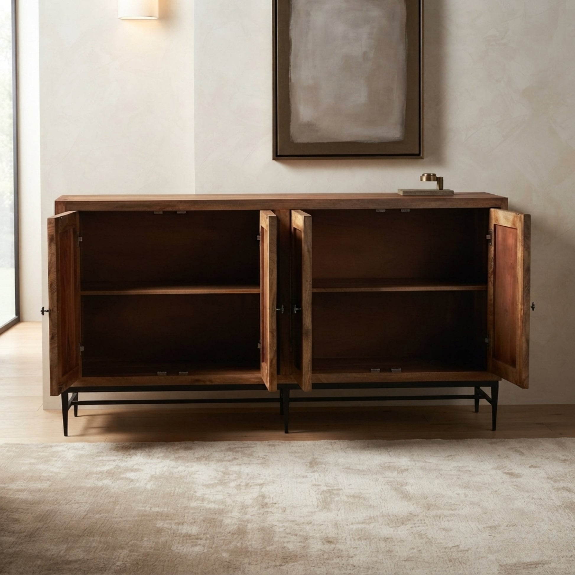 A rustic wooden sideboard cabinet with two open doors, showing its interior shelves, set against a neutral beige wall with a framed artwork and a small sink fixture on top.