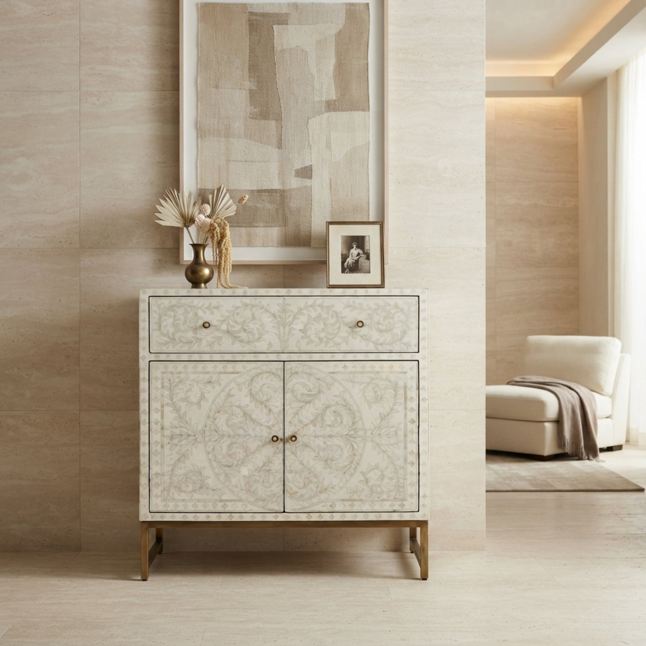 A cream-colored console cabinet with intricate shell-like floral patterns and brass legs, styled in a modern, warm-toned living room setting.