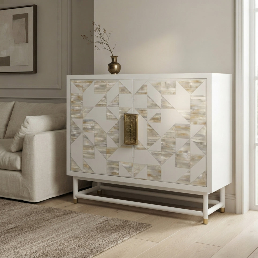 A white two-door cabinet with geometric shell-patterned doors and a central brass handle, styled in a modern living room next to a beige sofa.