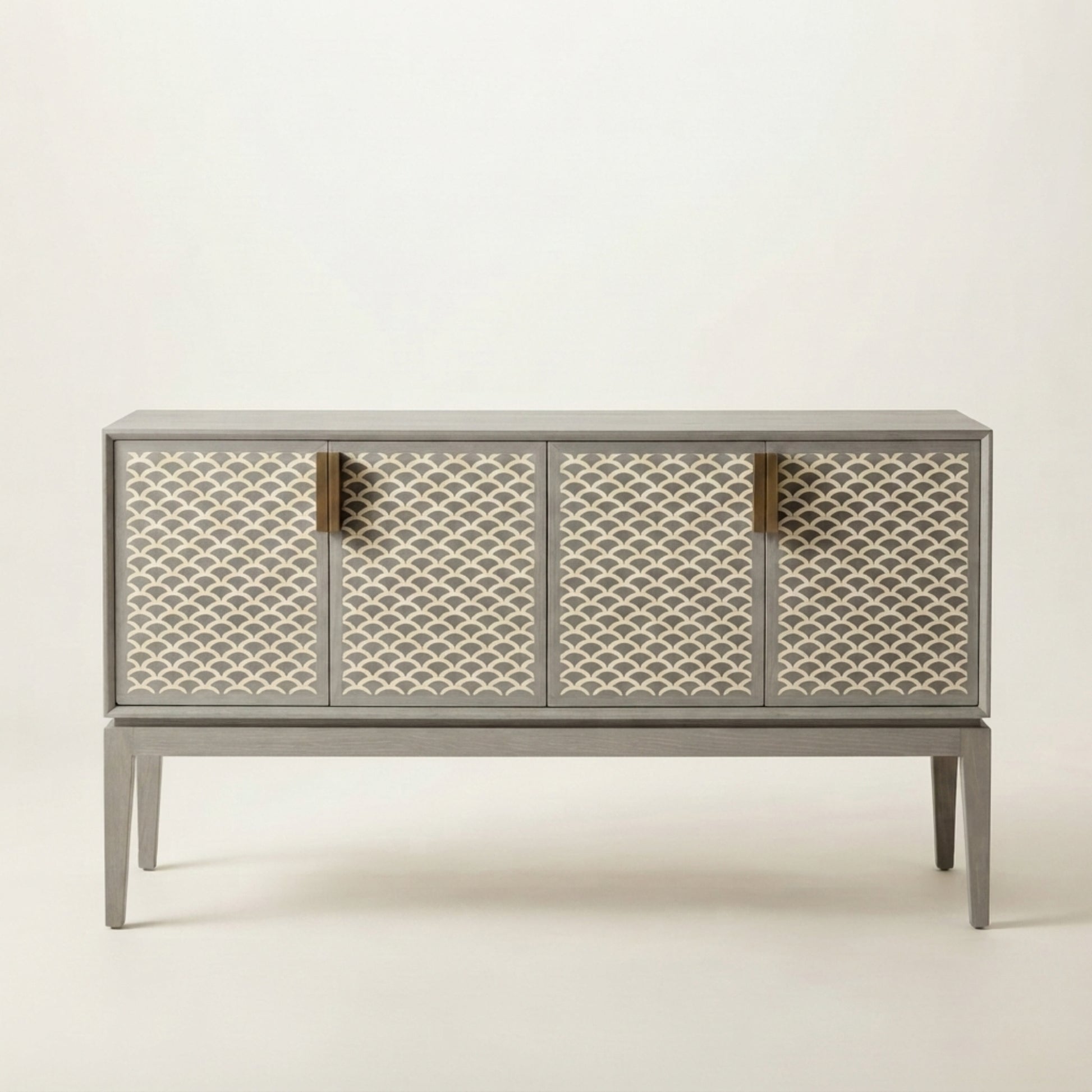 A modern sideboard cabinet with a gray frame and legs, featuring four doors decorated with a cream and gray geometric fish-scale pattern, accented by four vertical brass handles, set against a plain off-white background.