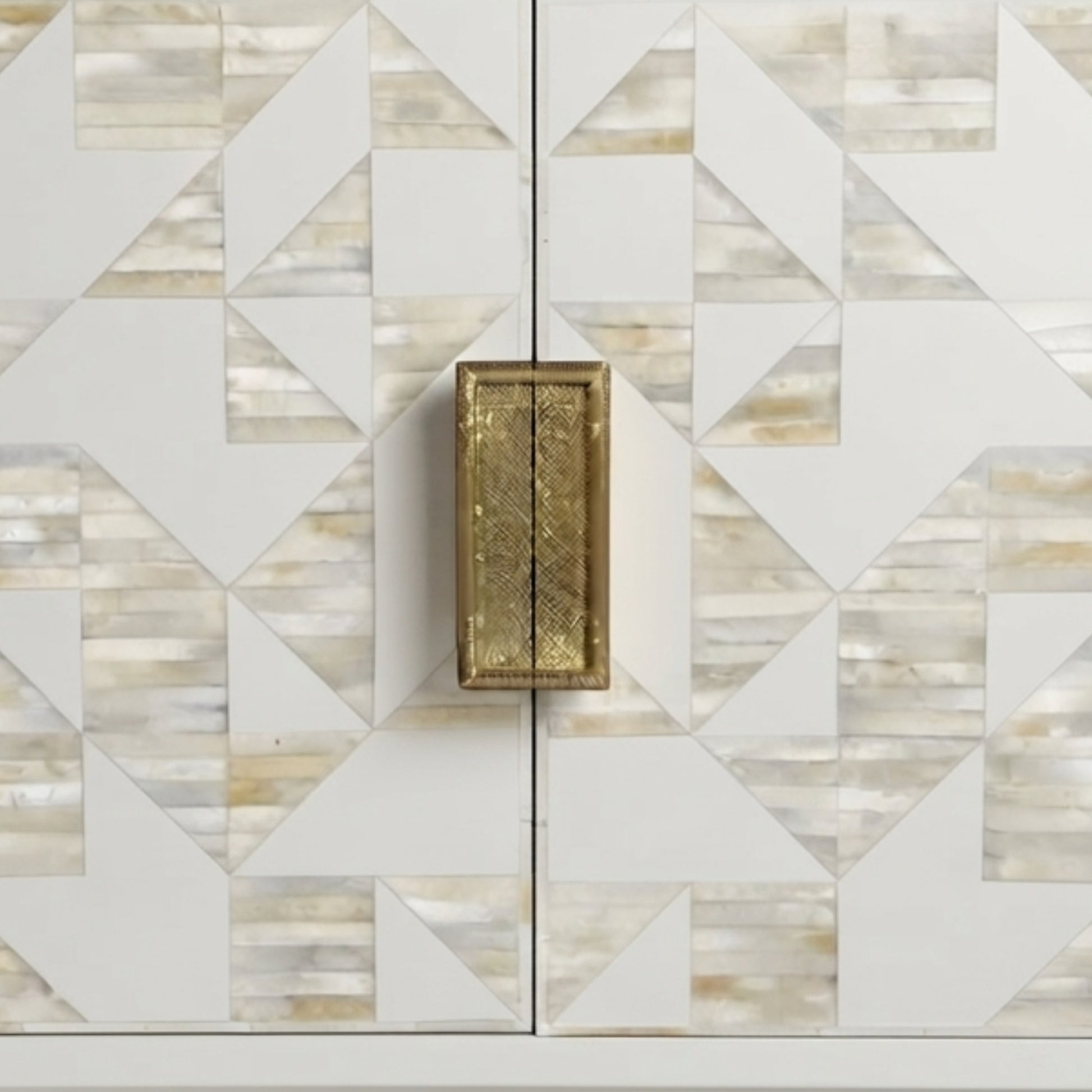 A close-up of a cabinet door featuring a geometric inlay pattern of white, beige, and light brown mother-of-pearl tiles, with a single rectangular brass handle centered on the split.