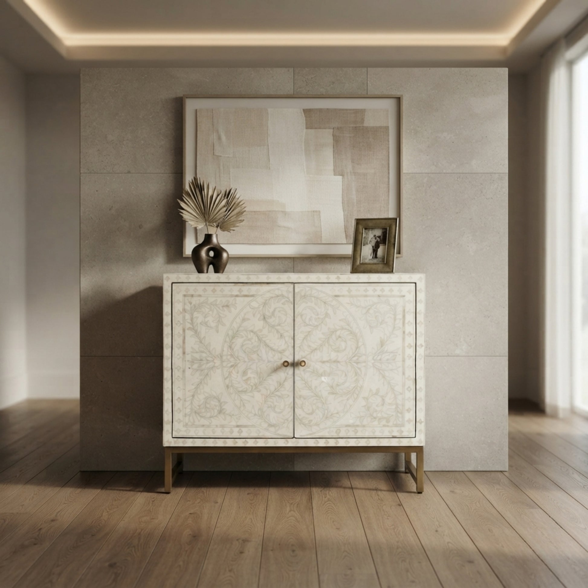 A cream-colored double-door cabinet with intricate bone inlay patterns and gold legs, styled in a modern interior setting with a framed abstract print and a small picture frame on top.