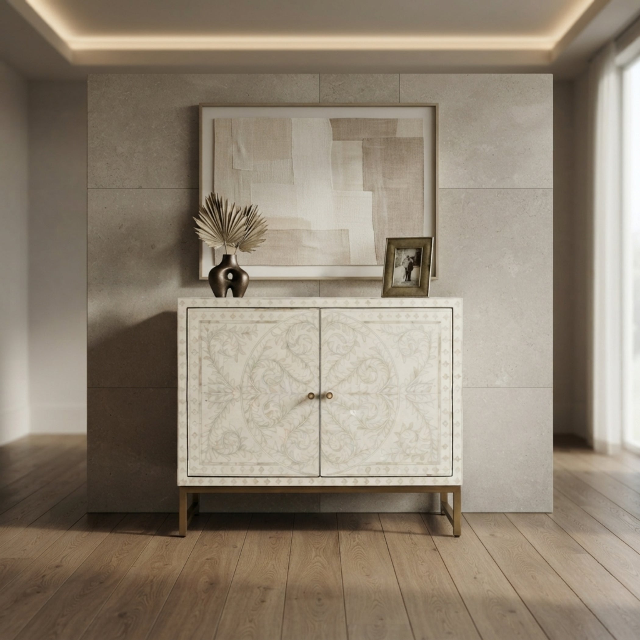 A cream-colored double-door cabinet with intricate bone inlay patterns and gold legs, styled in a modern interior setting with a framed abstract print and a small picture frame on top.
