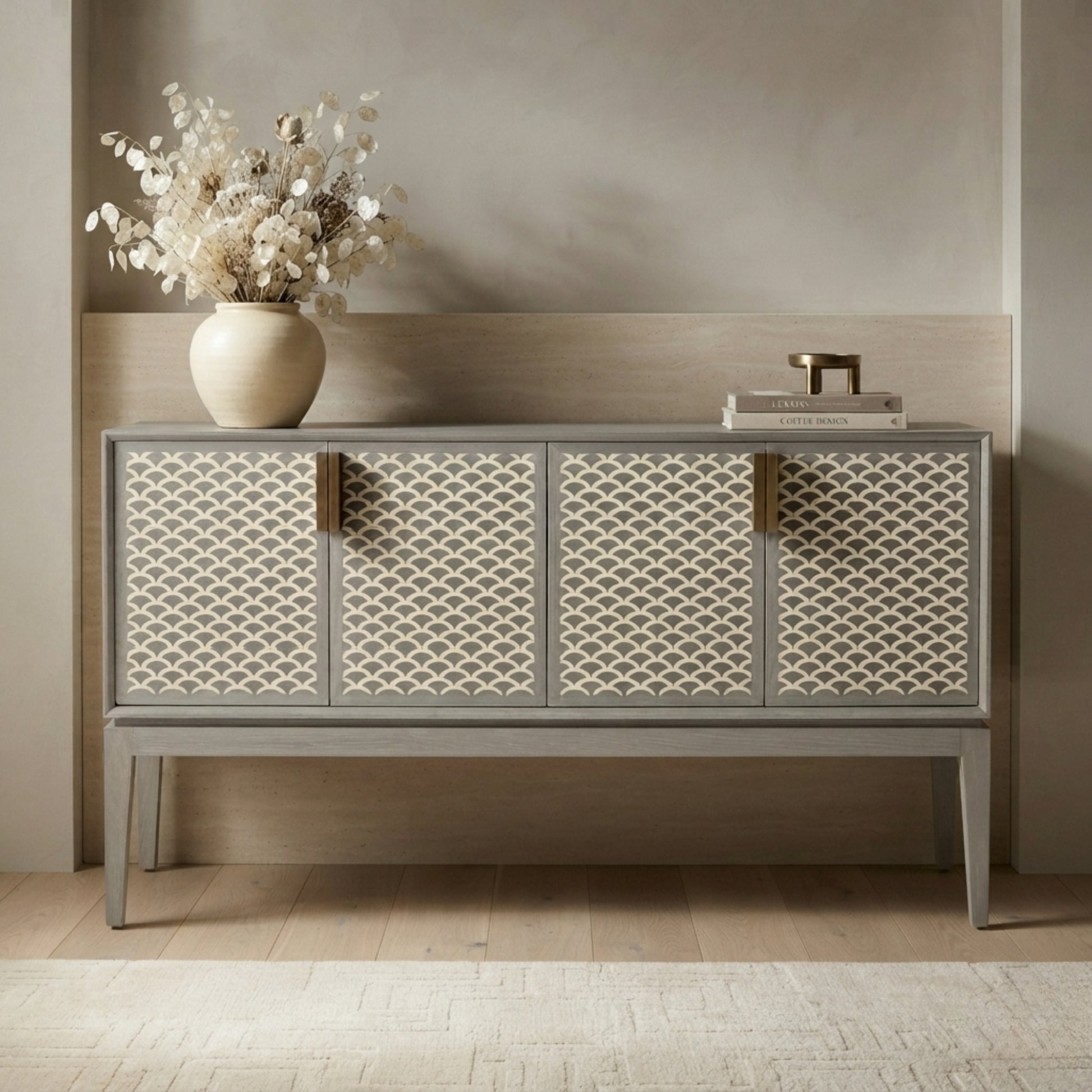 A gray sideboard cabinet with four patterned cream doors and brass handles, styled in a minimalist room with a vase of dried flowers on top.