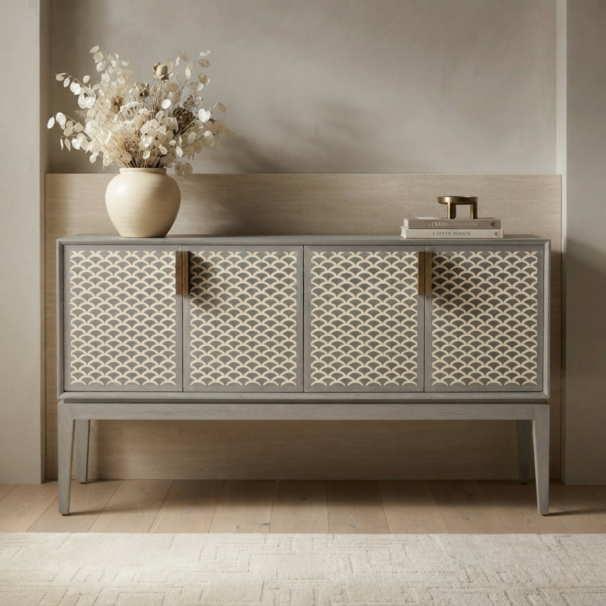 A gray sideboard cabinet with four patterned cream doors and brass handles, styled in a minimalist room with a vase of dried flowers on top.
