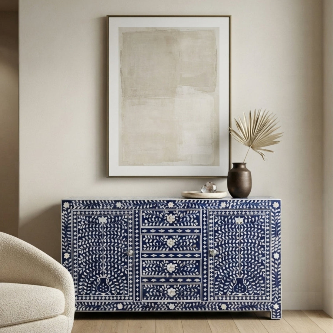 A blue and white patterned sideboard cabinet in a styled living room setting, featuring a cream armchair, a framed abstract print, and a small vase with dried leaves on top.