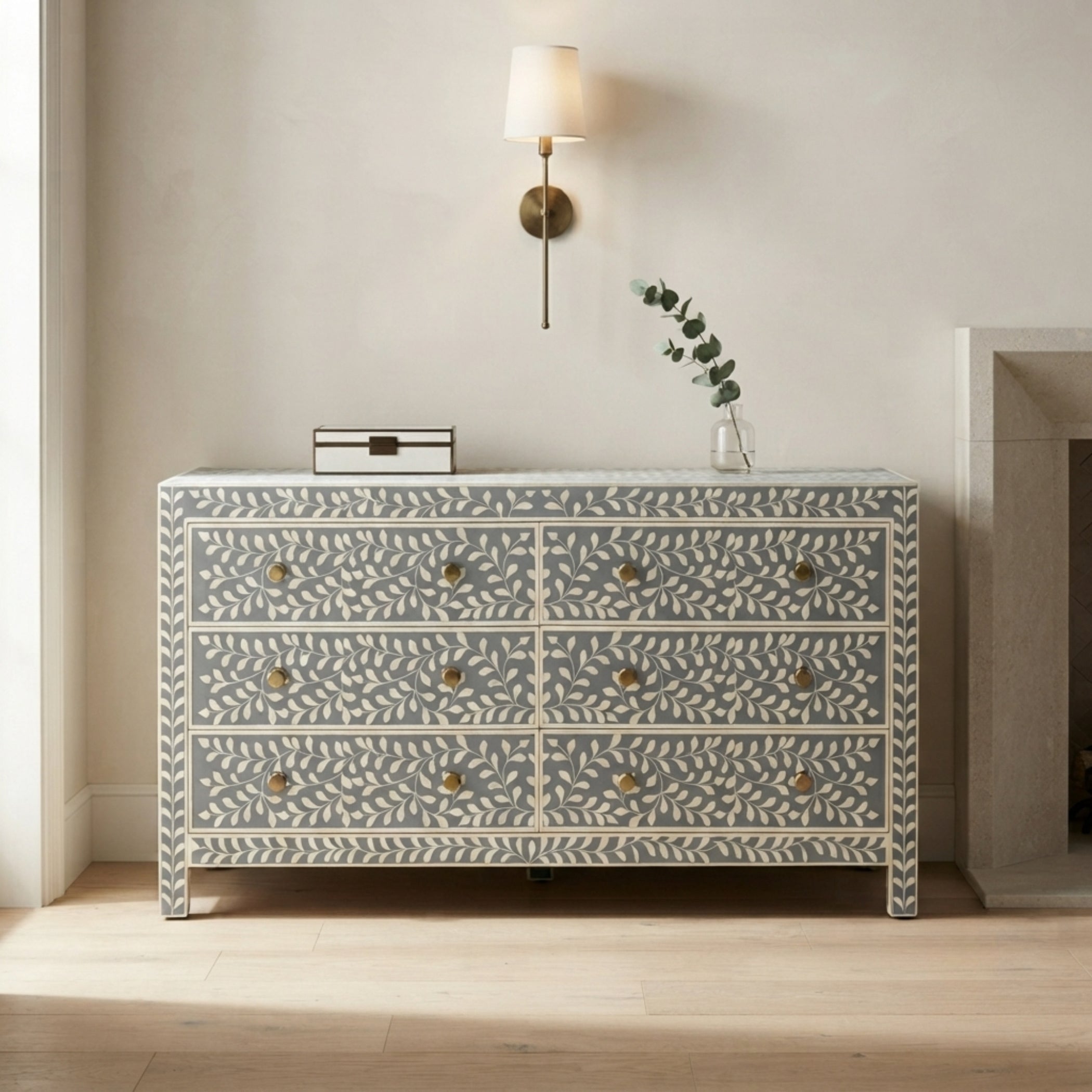 A six-drawer chest of drawers with a blue-gray and cream bone-inlay pattern and brass knobs, styled in a minimalist room setting with a wall sconce and a small vase on top.