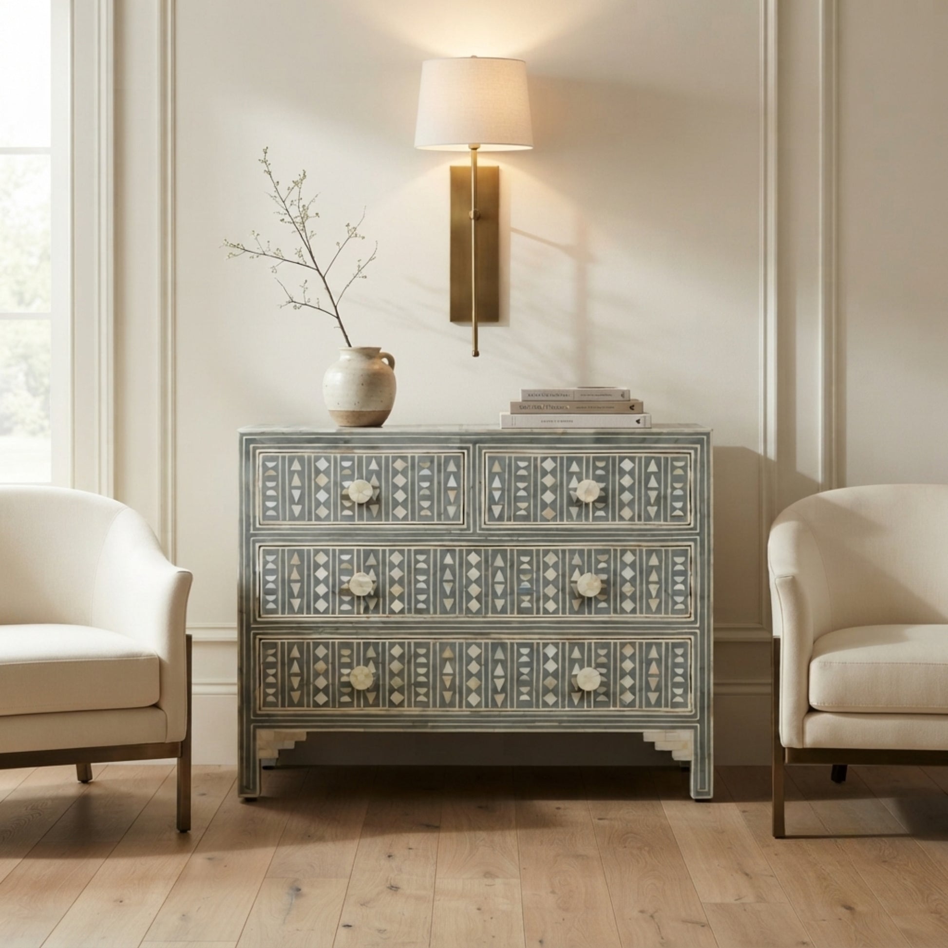 A decorative chest of drawers with intricate geometric patterns in gray and cream, styled in a bright living room between two cream armchairs under a wall sconce.