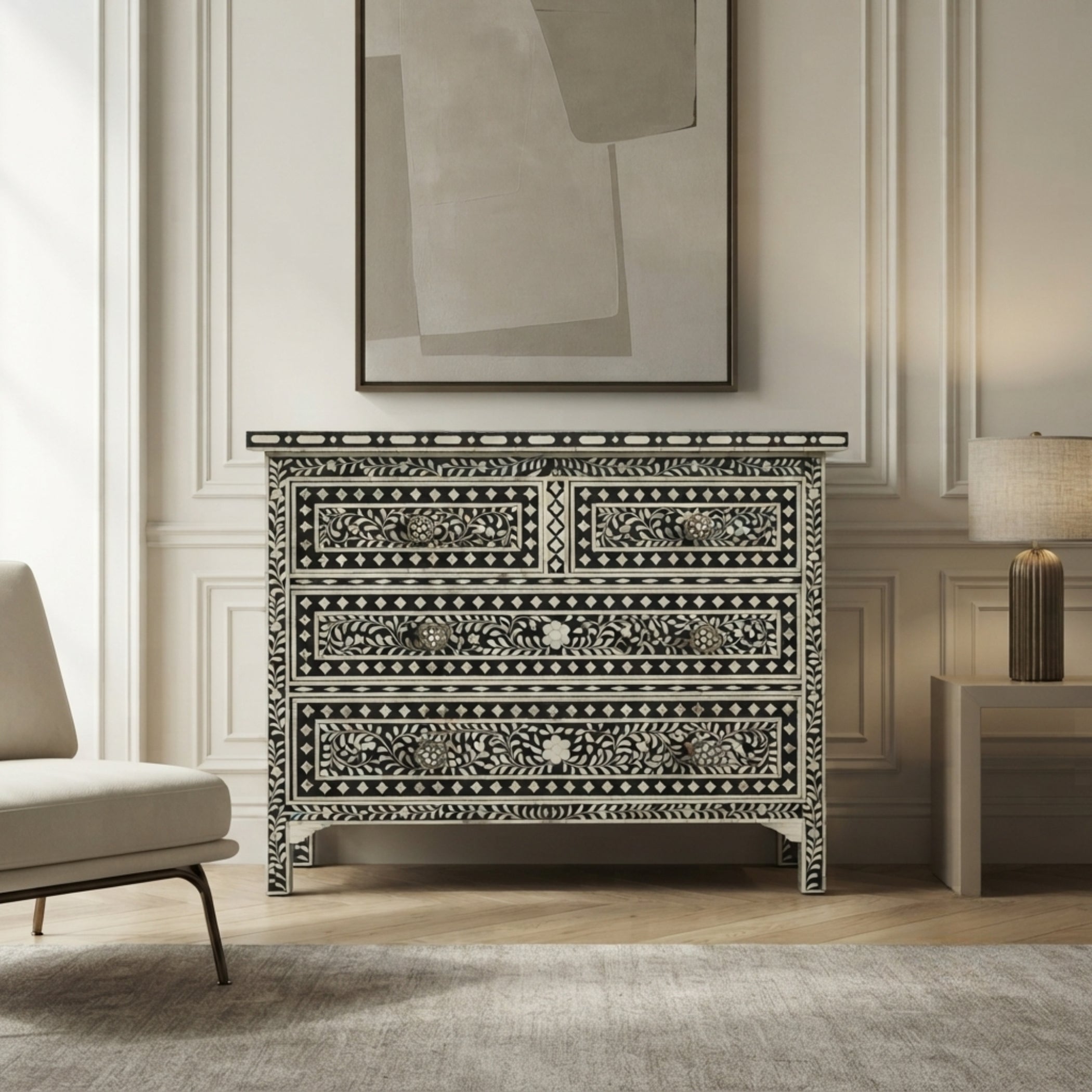A black and white bone-inlay dresser with three drawers stands in a styled room setting with a light gray armchair, a side table with a lamp, and a framed abstract print on the wall.