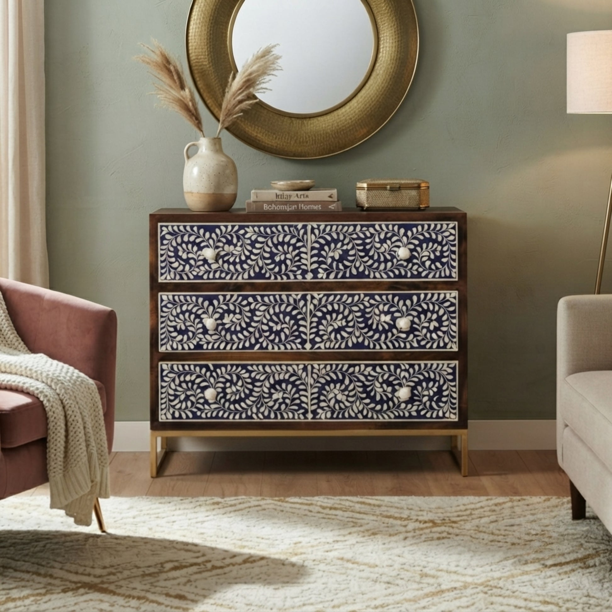 A three-drawer chest of drawers with navy blue and white floral patterns and gold legs, styled in a living room setting with a round mirror, pampas grass, and armchairs.