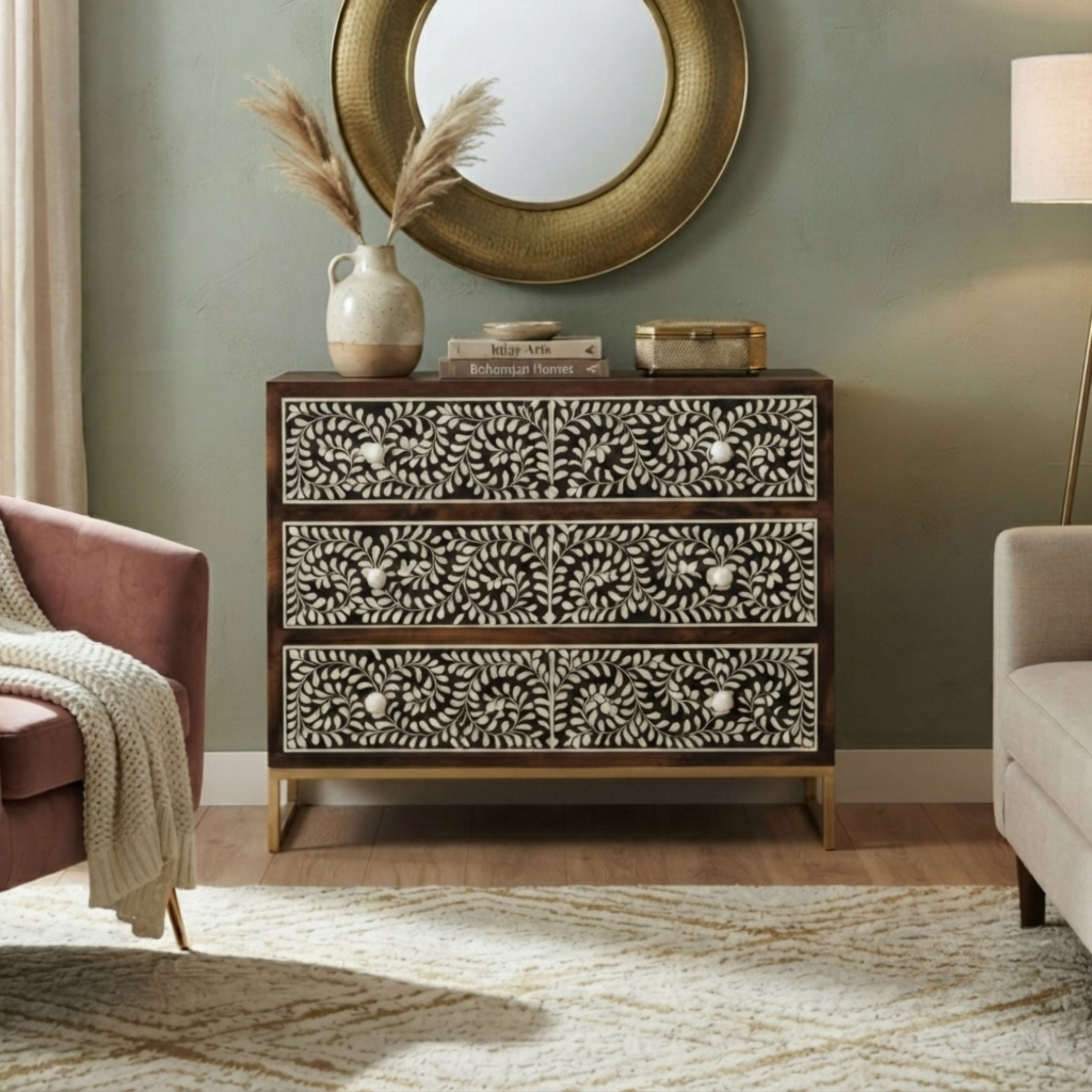 A three-drawer chest of drawers with black and white floral bone-style drawer fronts and gold-tone legs, styled in a living room setting with a mirror, lamp, and armchair.
