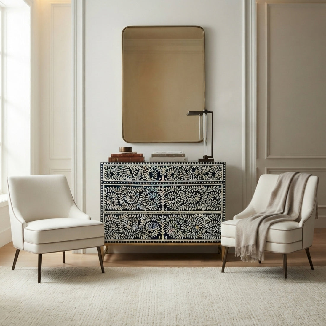 A black and white bone-inlay console table with a matching rectangular mirror, styled in a bright entryway with two cream armchairs and a draped throw blanket.