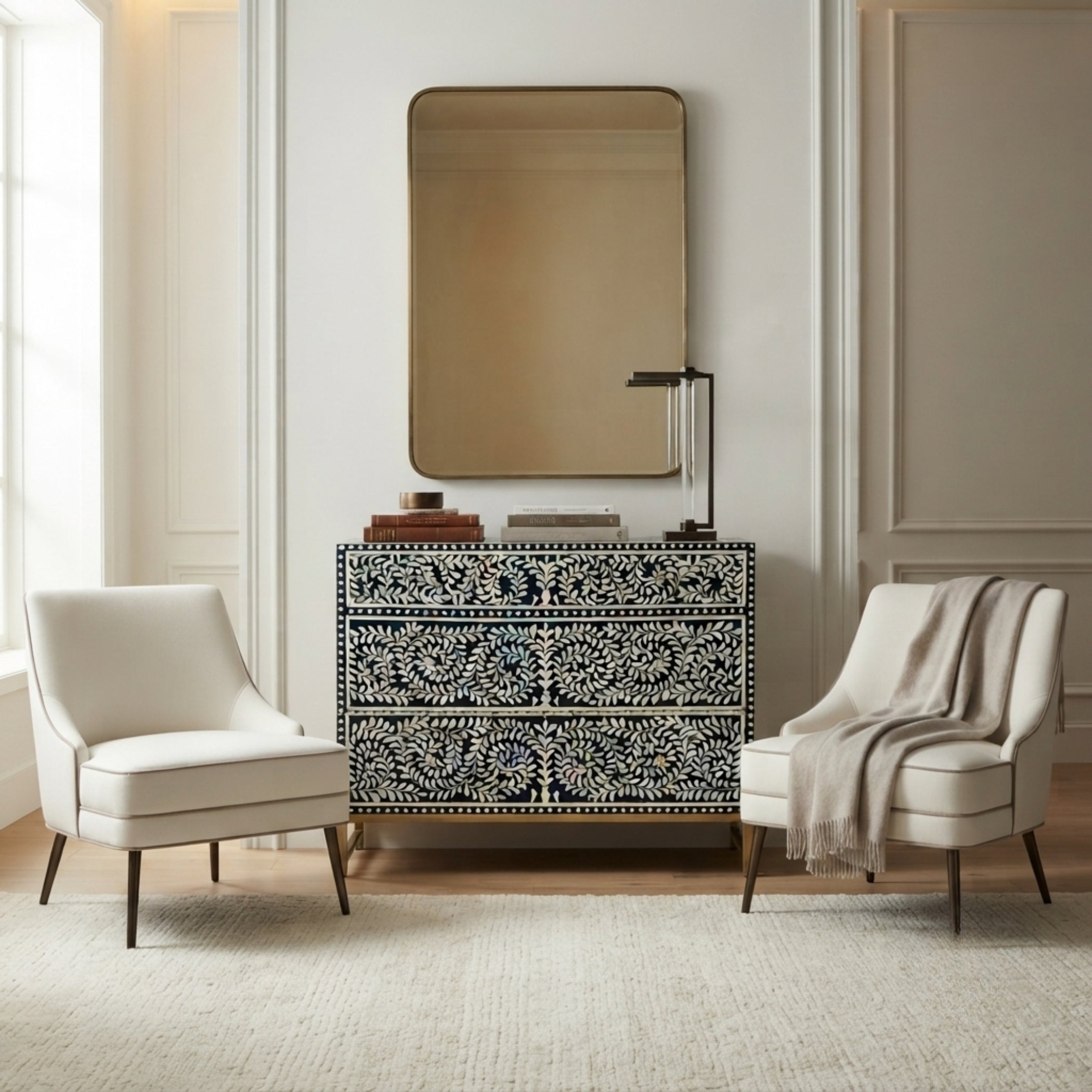 A black and white bone-inlay console table with a matching rectangular mirror, styled in a bright entryway with two cream armchairs and a draped throw blanket.