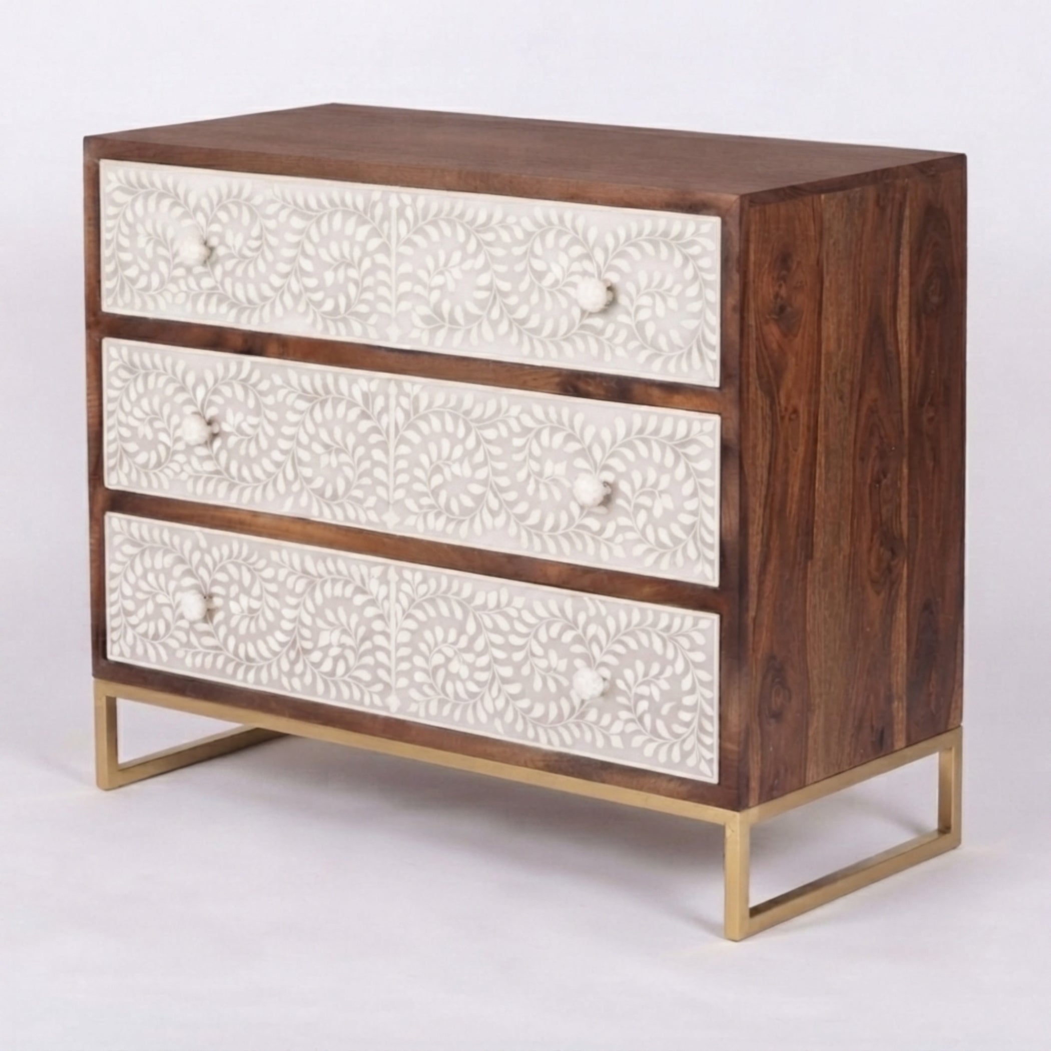 A three-drawer wooden chest of drawers with carved off-white drawer fronts, a dark brown wood body, and a gold metal frame base, shown on a light gray studio background.
