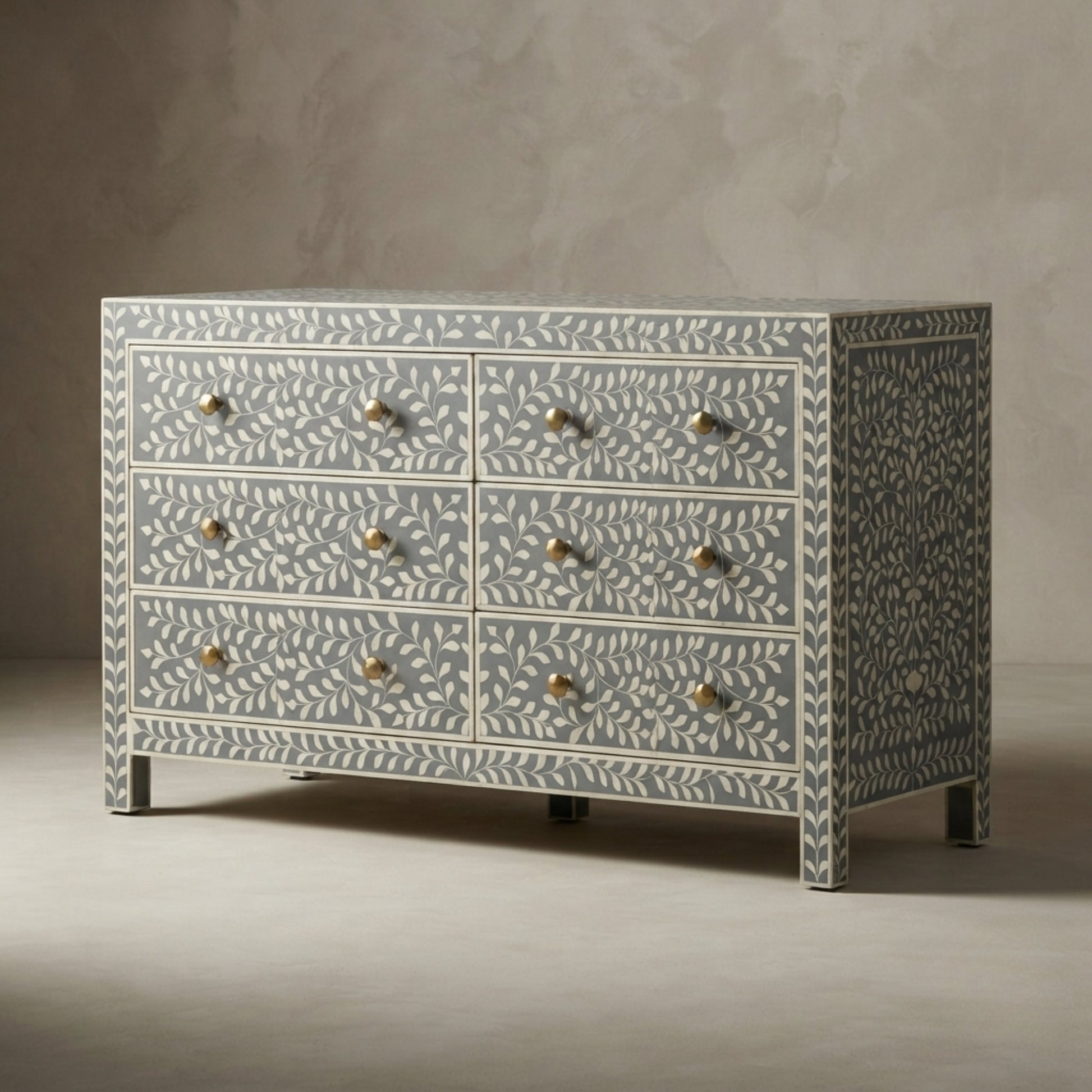 A six-drawer dresser with a light blue and white bone-inlay pattern and brass knobs, set against a textured gray studio background.
