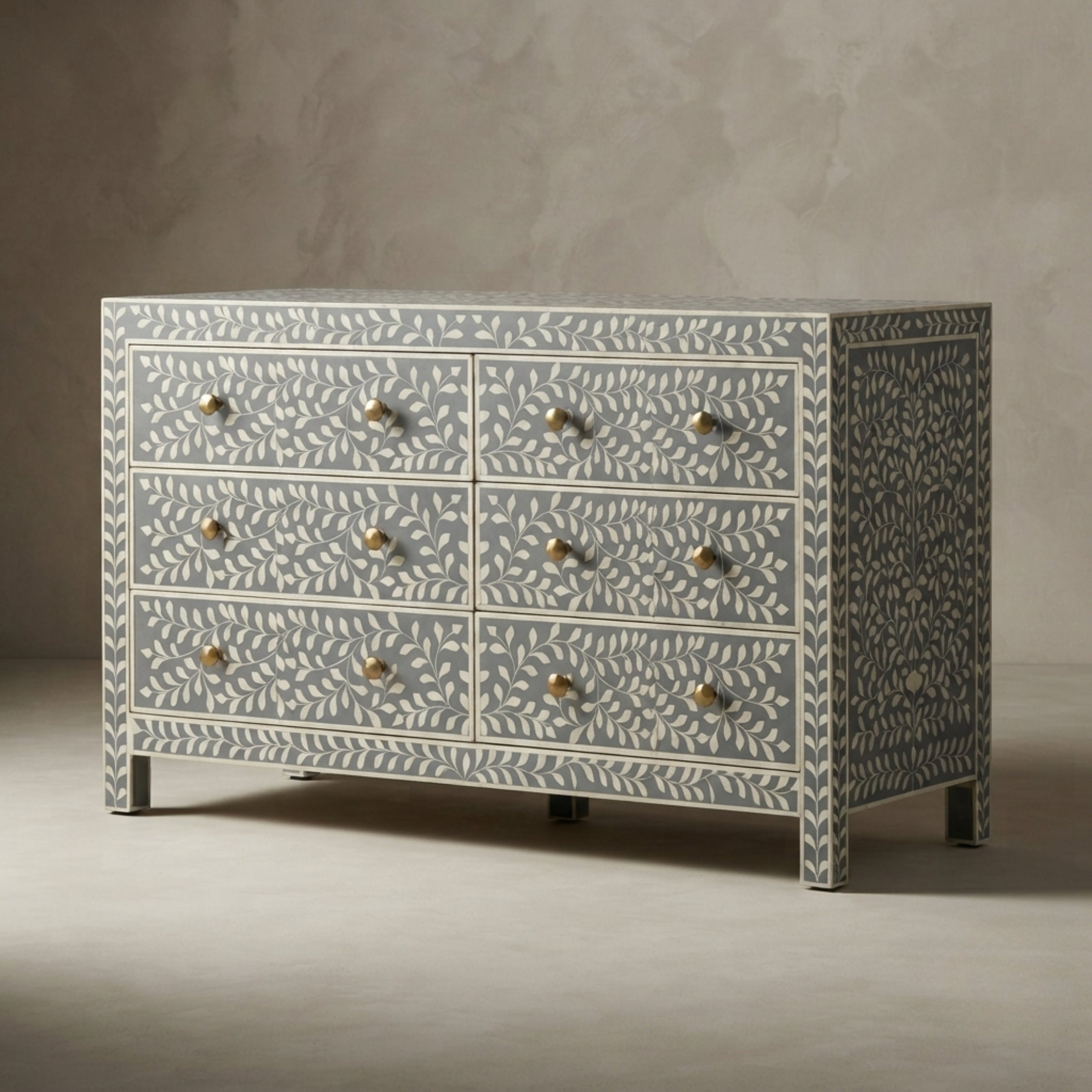 A six-drawer dresser with a light blue and white bone-inlay pattern and brass knobs, set against a textured gray studio background.