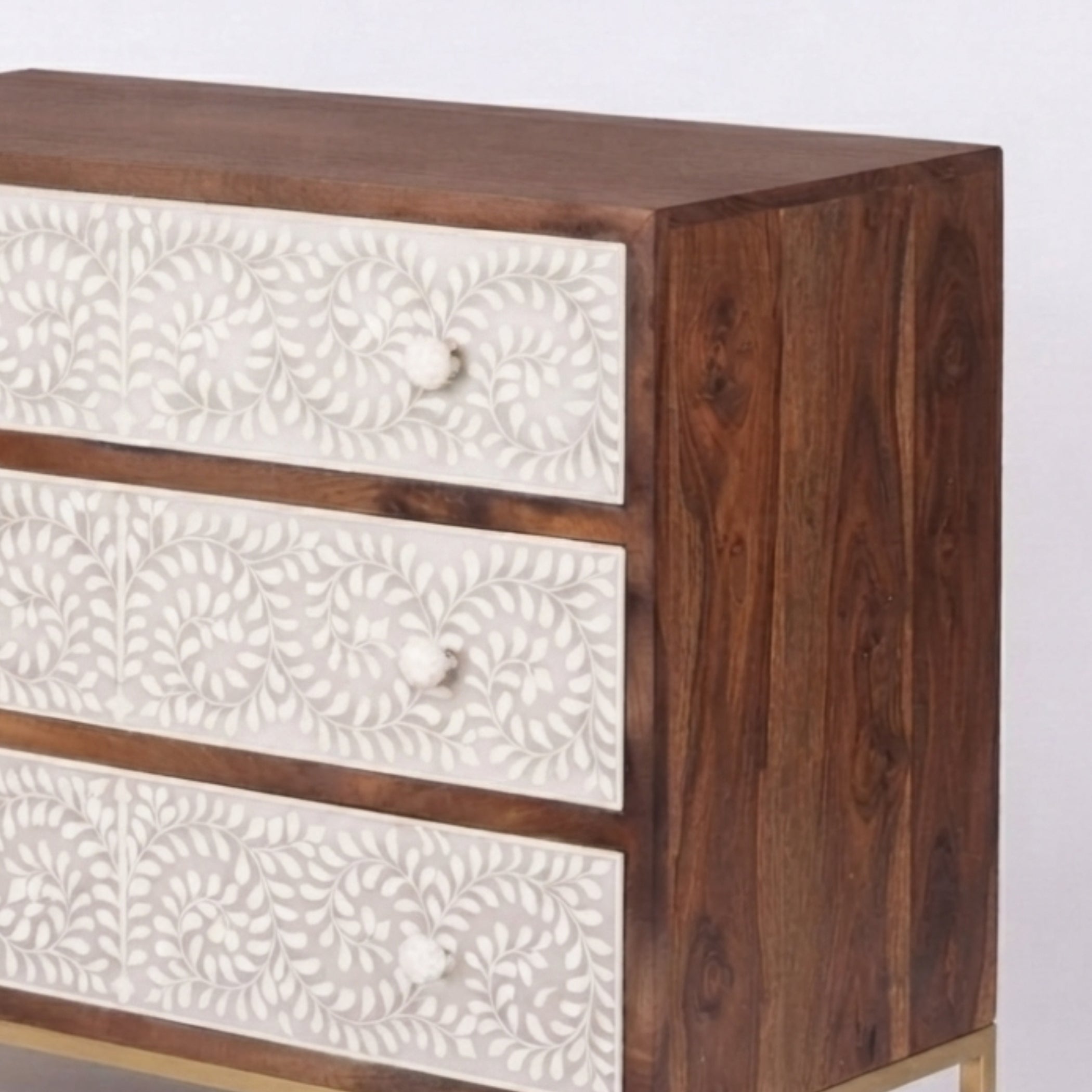 A close-up of a wooden chest of drawers with three drawers featuring carved white floral panels and small round knobs, set against a plain light background.