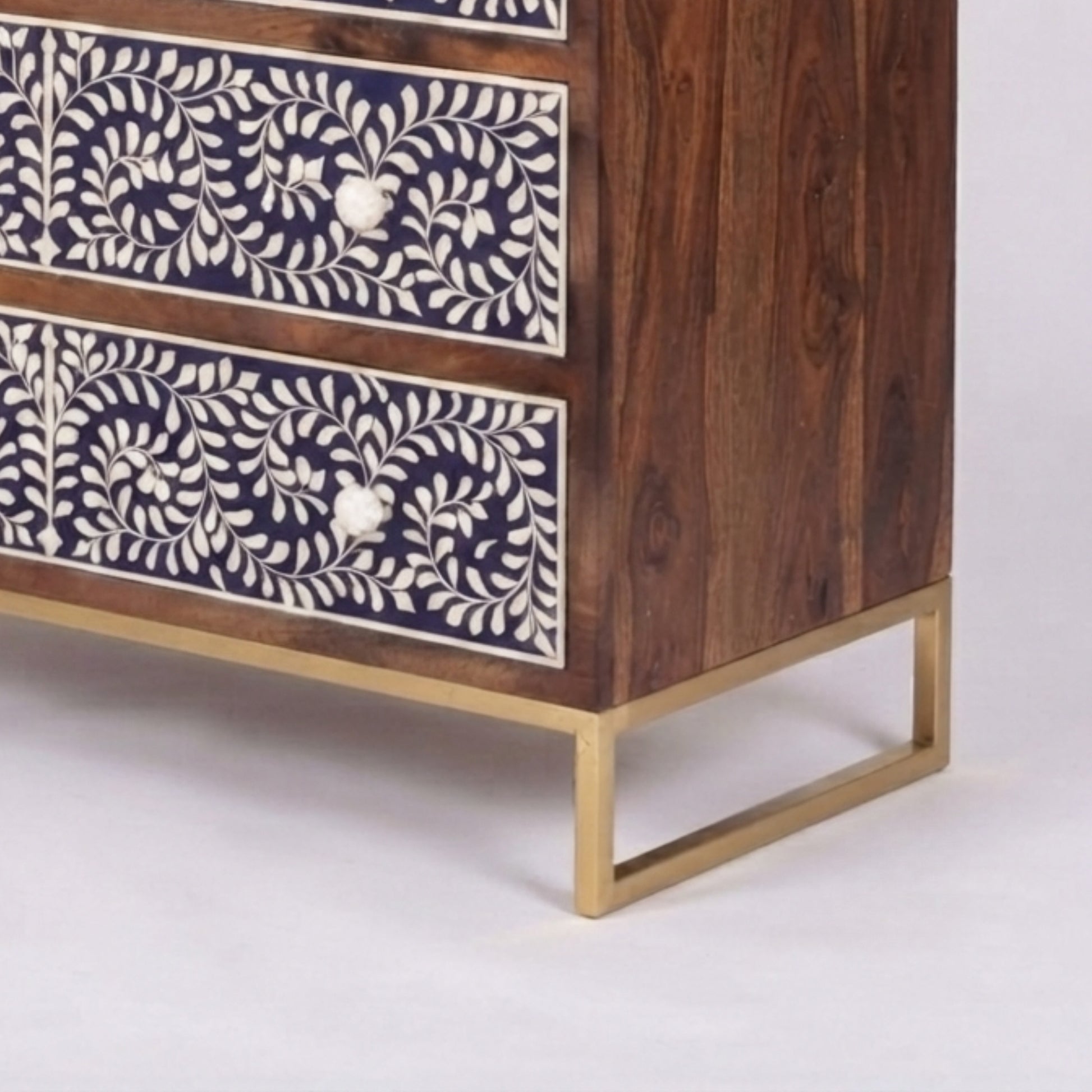 A close-up of a wooden chest of drawers with dark brown wood grain, cream bone-inlay floral patterns on the drawer fronts, and a gold metal frame base, shown on a light gray background.