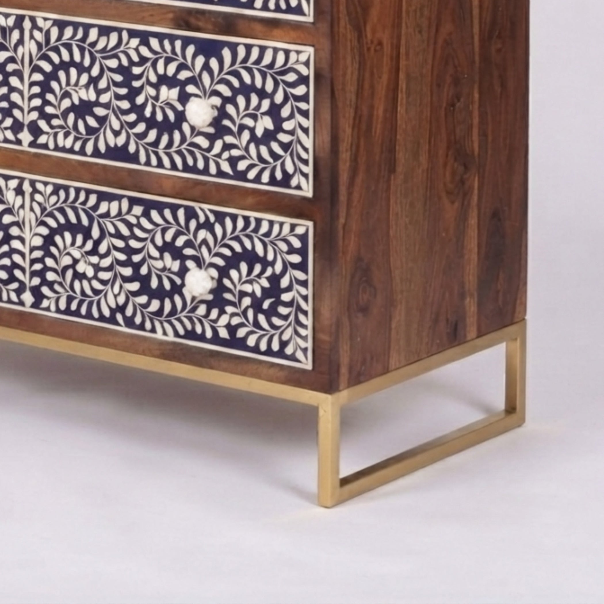 A close-up of a wooden chest of drawers with dark brown wood grain, cream bone-inlay floral patterns on the drawer fronts, and a gold metal frame base, shown on a light gray background.
