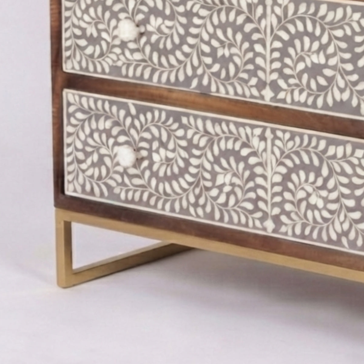 Close-up of a wooden chest of drawers with two drawers featuring intricate bone-inlay floral patterns in cream and brown, with a gold metal leg frame, on a light gray background.