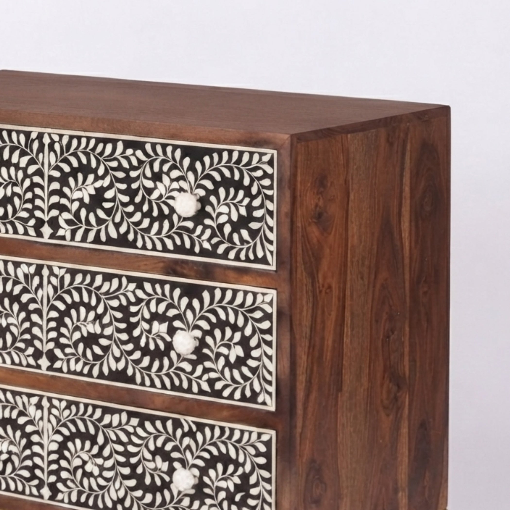 A close-up of a wooden chest of drawers with three visible drawers, featuring dark brown wood grain and intricate white bone or mother-of-pearl floral inlay on the drawer fronts.