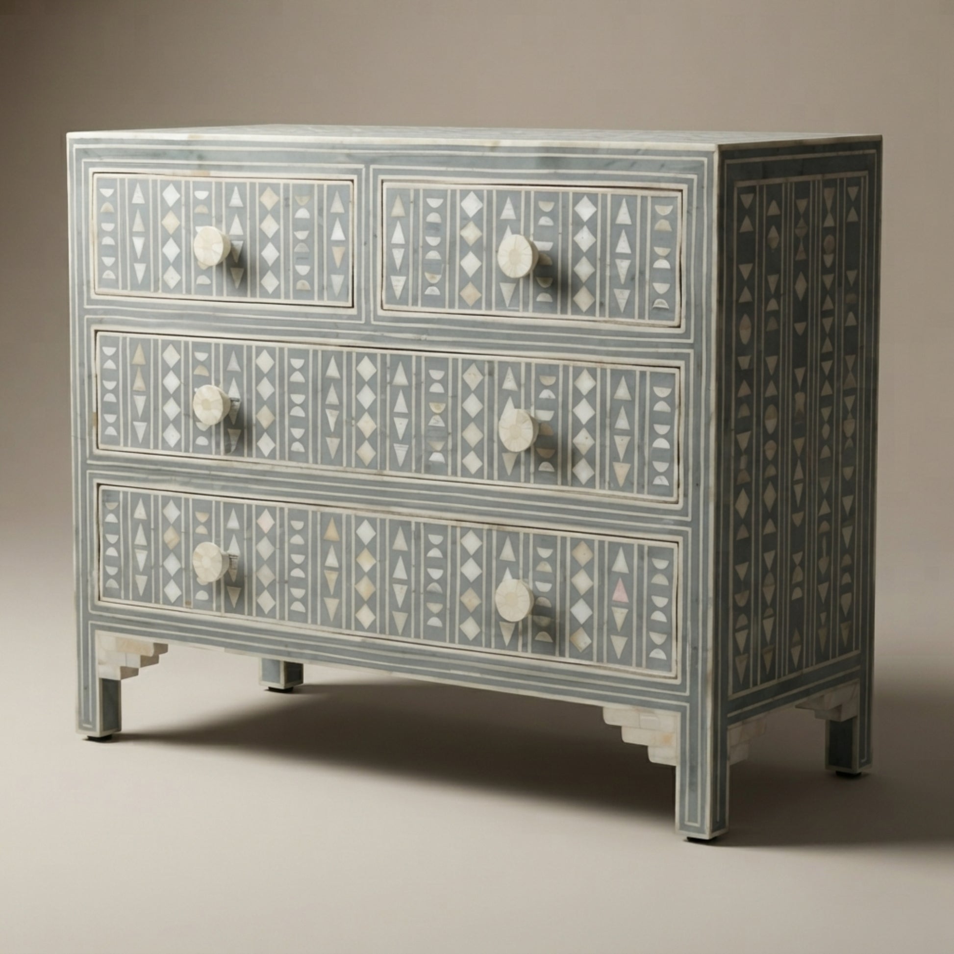 A three-drawer chest of drawers with a gray painted finish and intricate white bone or mother-of-pearl geometric inlay patterns, featuring round white knobs and tiered legs.