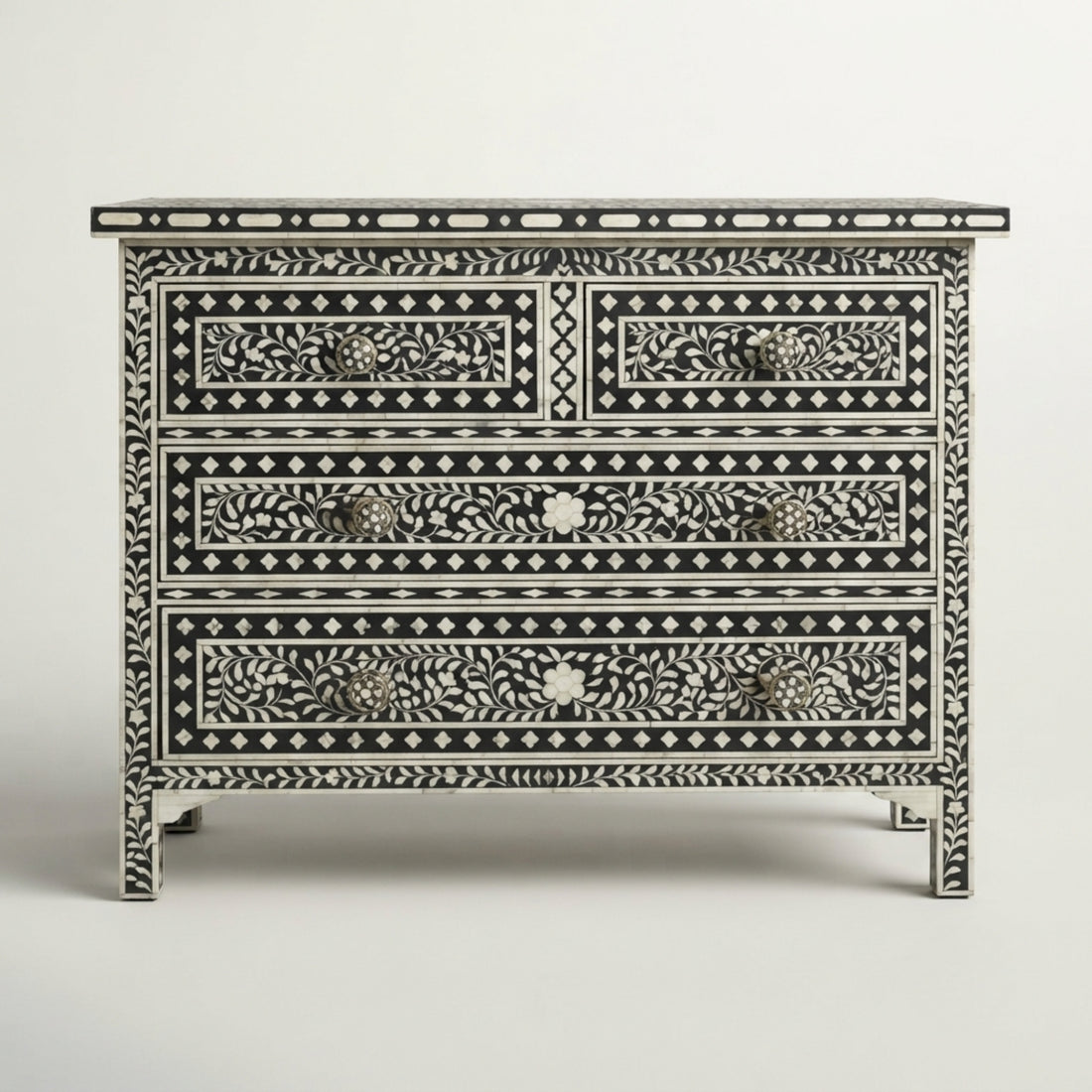 A black and white bone-inlay dresser with three drawers stands in a styled room setting with a light gray armchair, a side table with a lamp, and a framed abstract print on the wall.