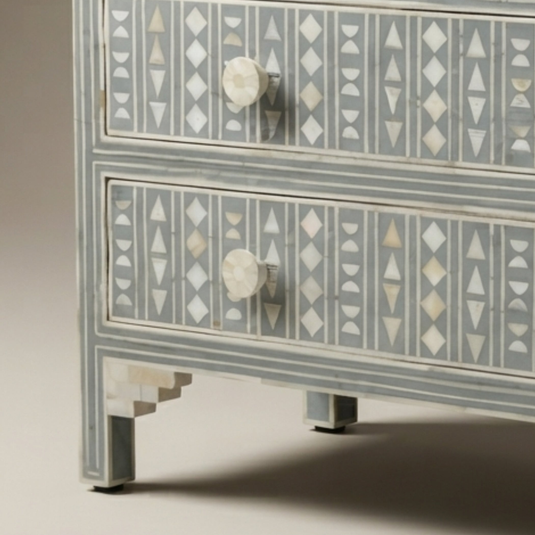 A close-up of a two-drawer chest of drawers with a light gray painted finish and intricate white and beige bone-inlay geometric patterns on the drawer fronts, featuring round white knobs and decorative legs.