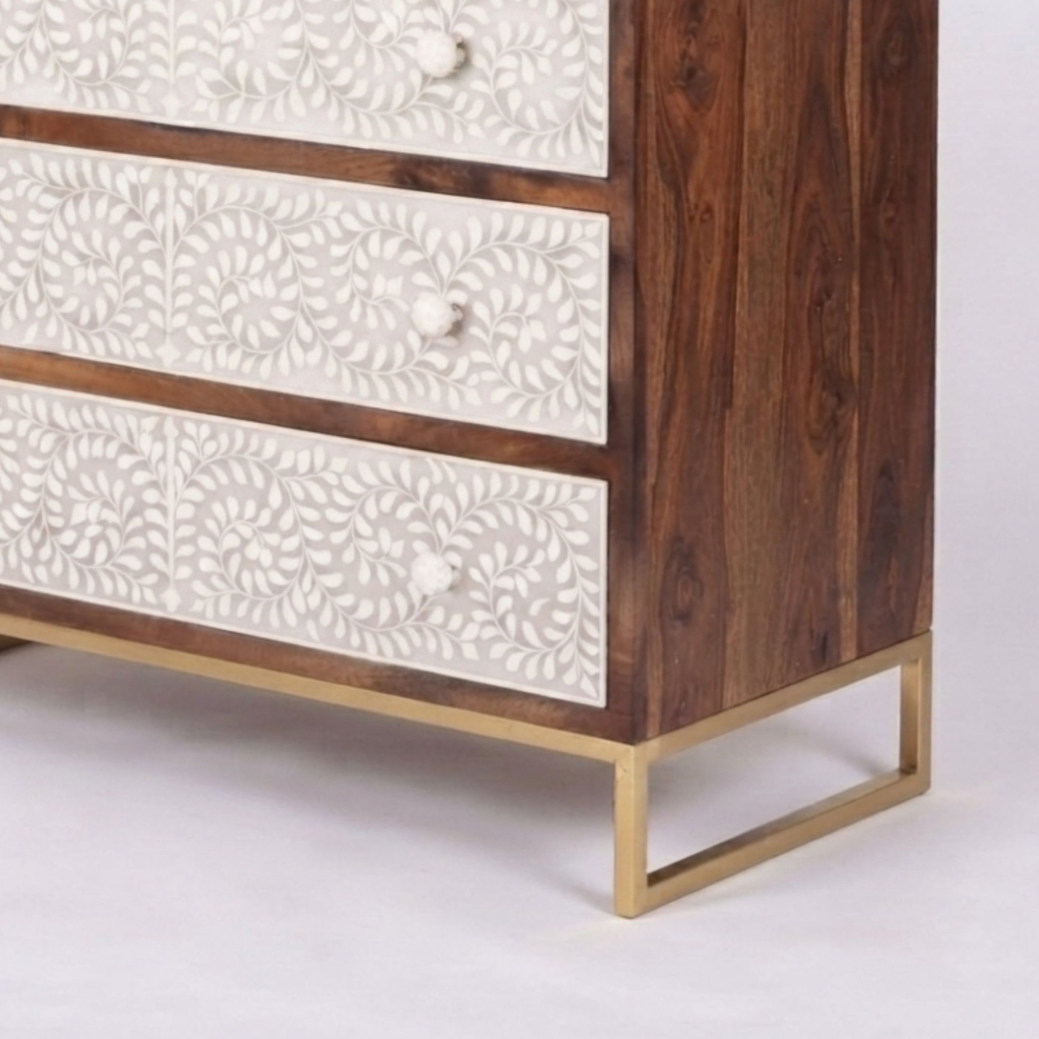 A close-up of a wooden chest of drawers with three cream-colored drawers featuring intricate white floral bone inlay patterns, supported by a gold-finished metal frame.