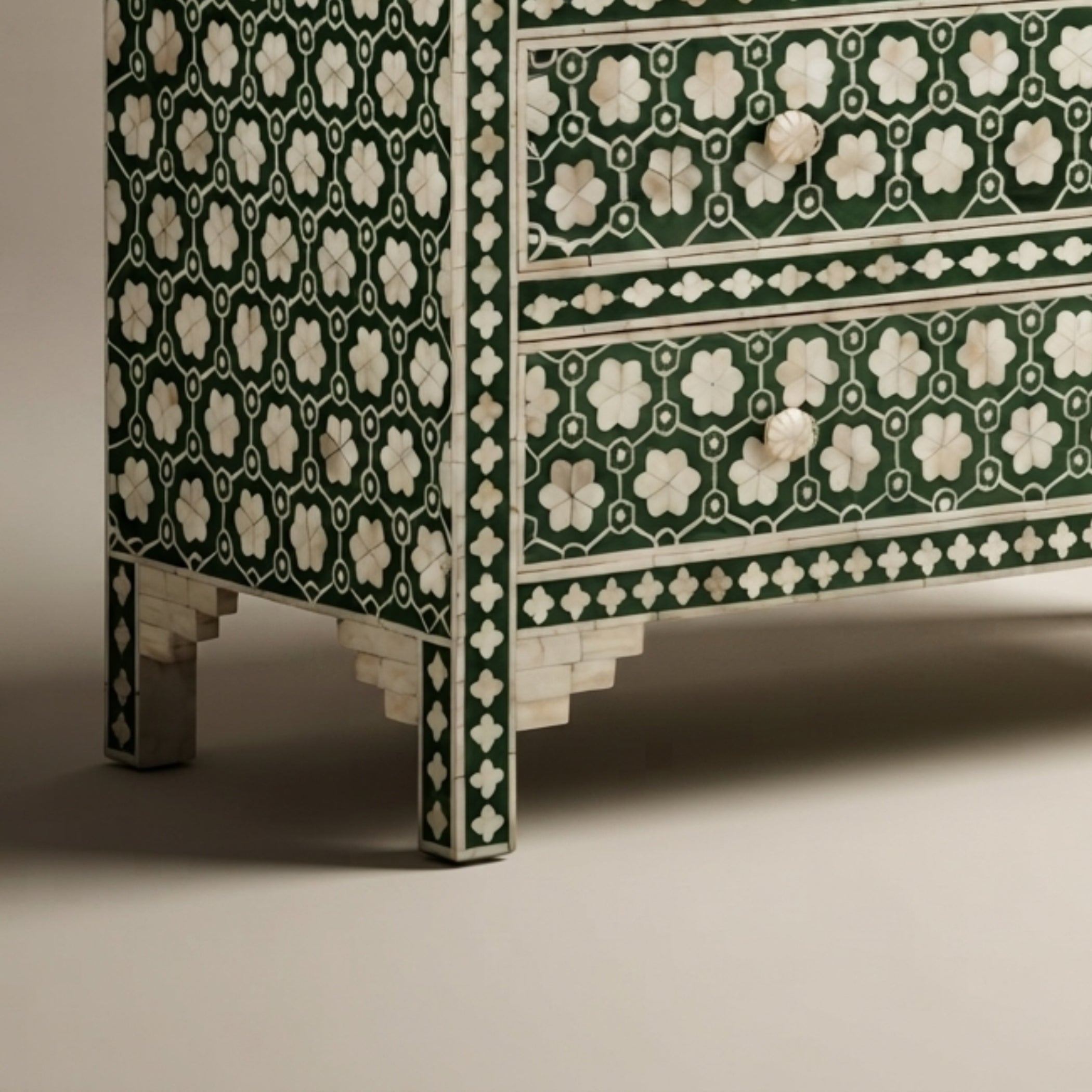 Close-up of a decorative chest of drawers with a dark green and white floral inlay pattern, featuring three drawers and ornate legs on a neutral studio background.
