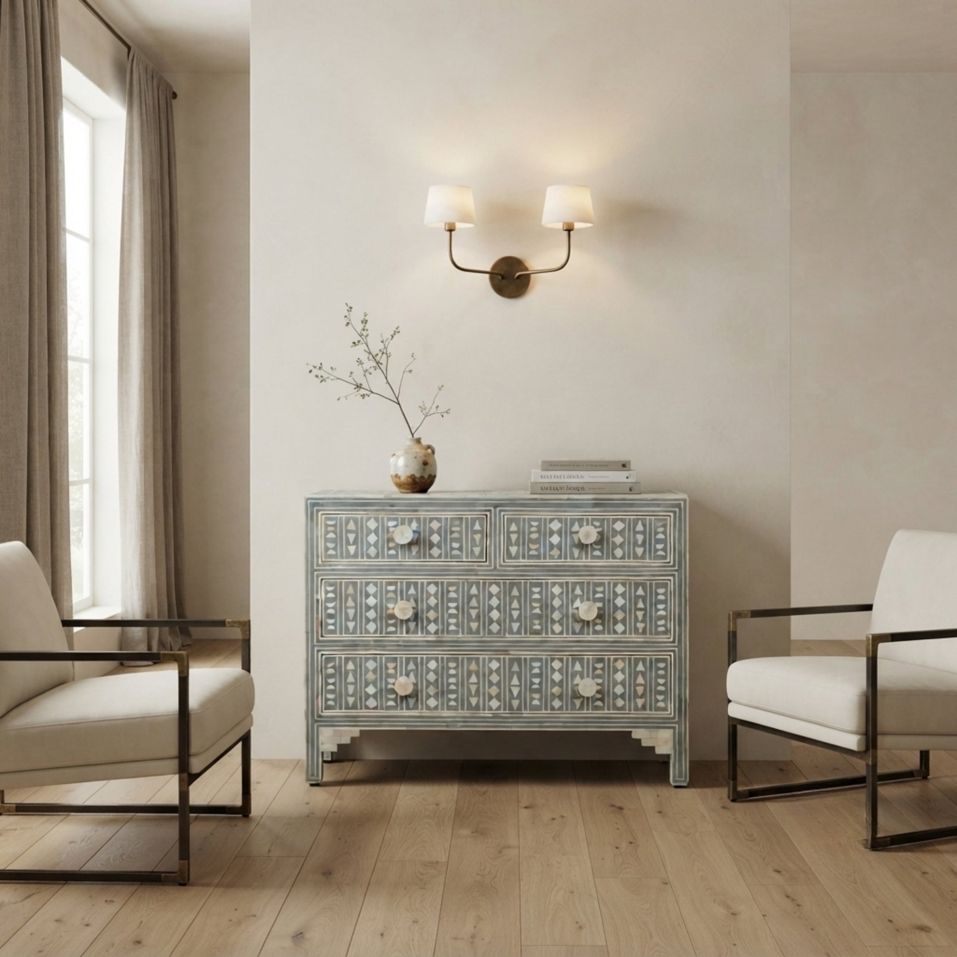 A light blue and white patterned three-drawer chest of drawers stands in a styled room with light wood floors, two cream armchairs, a wall-mounted double sconce, and a vase with branches on top.