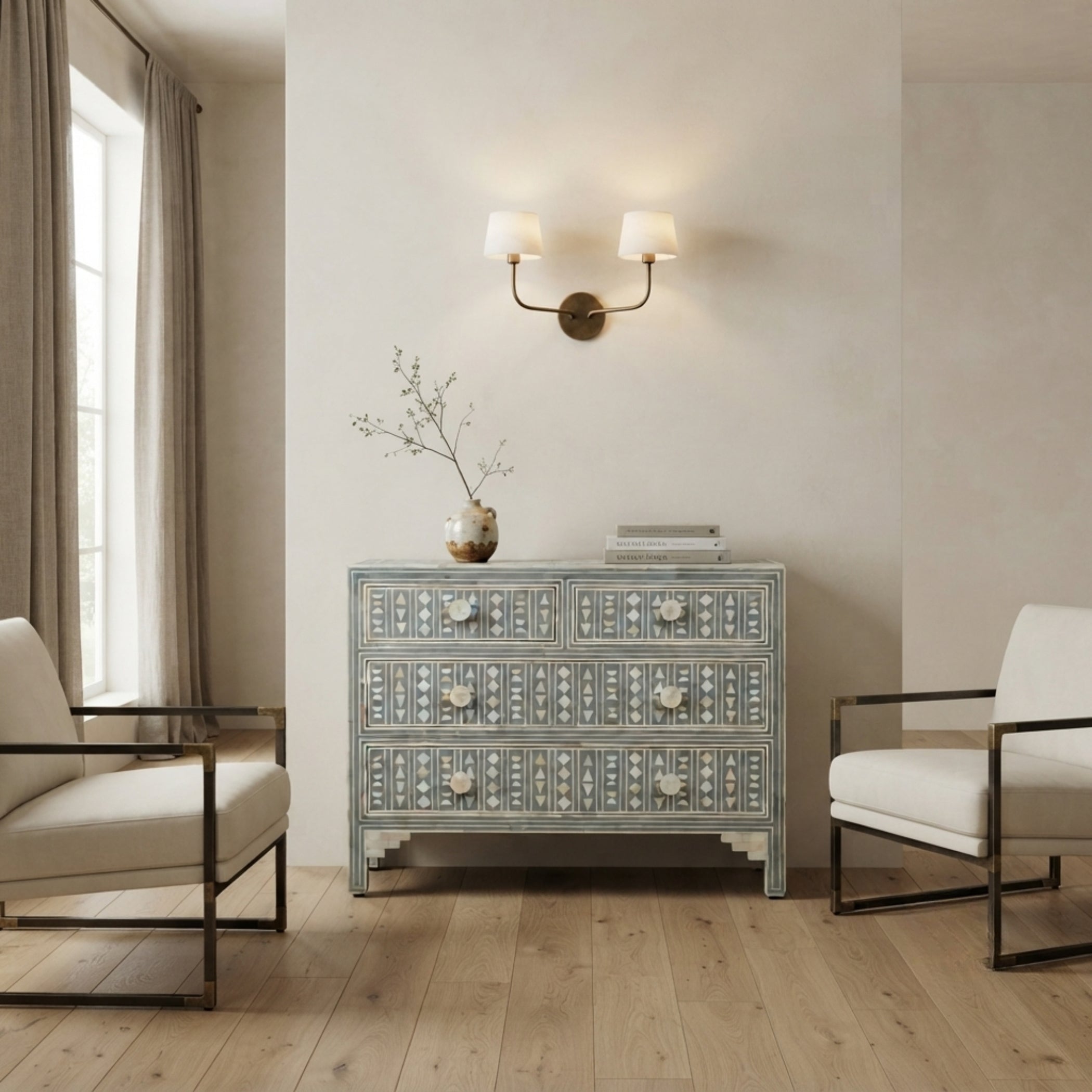A light blue and white patterned three-drawer chest of drawers stands in a styled room with light wood floors, two cream armchairs, a wall-mounted double sconce, and a vase with branches on top.