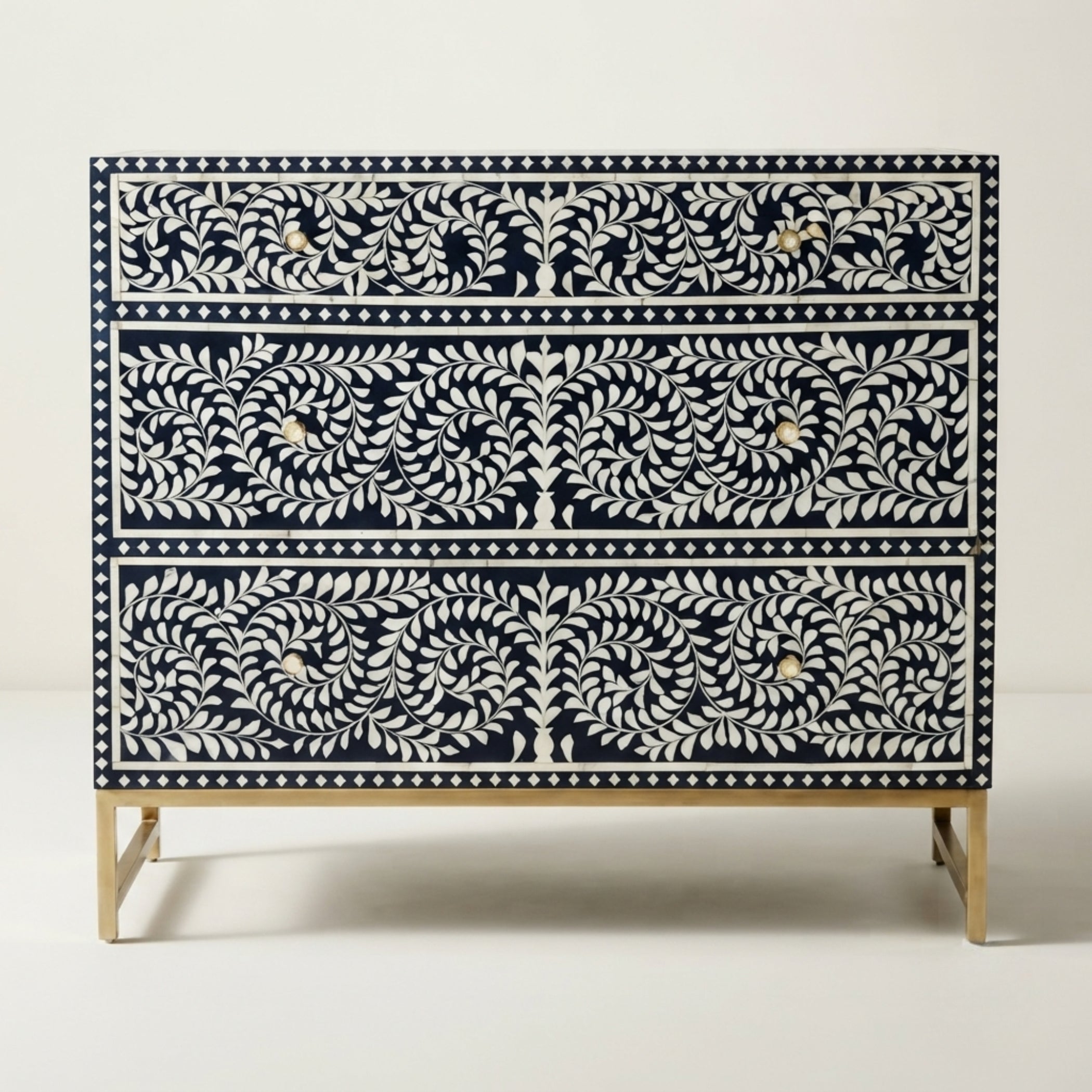 A three-drawer chest of drawers featuring an intricate bone-inlay pattern of white leafy vines and birds on a dark navy blue background, supported by a light wood base.