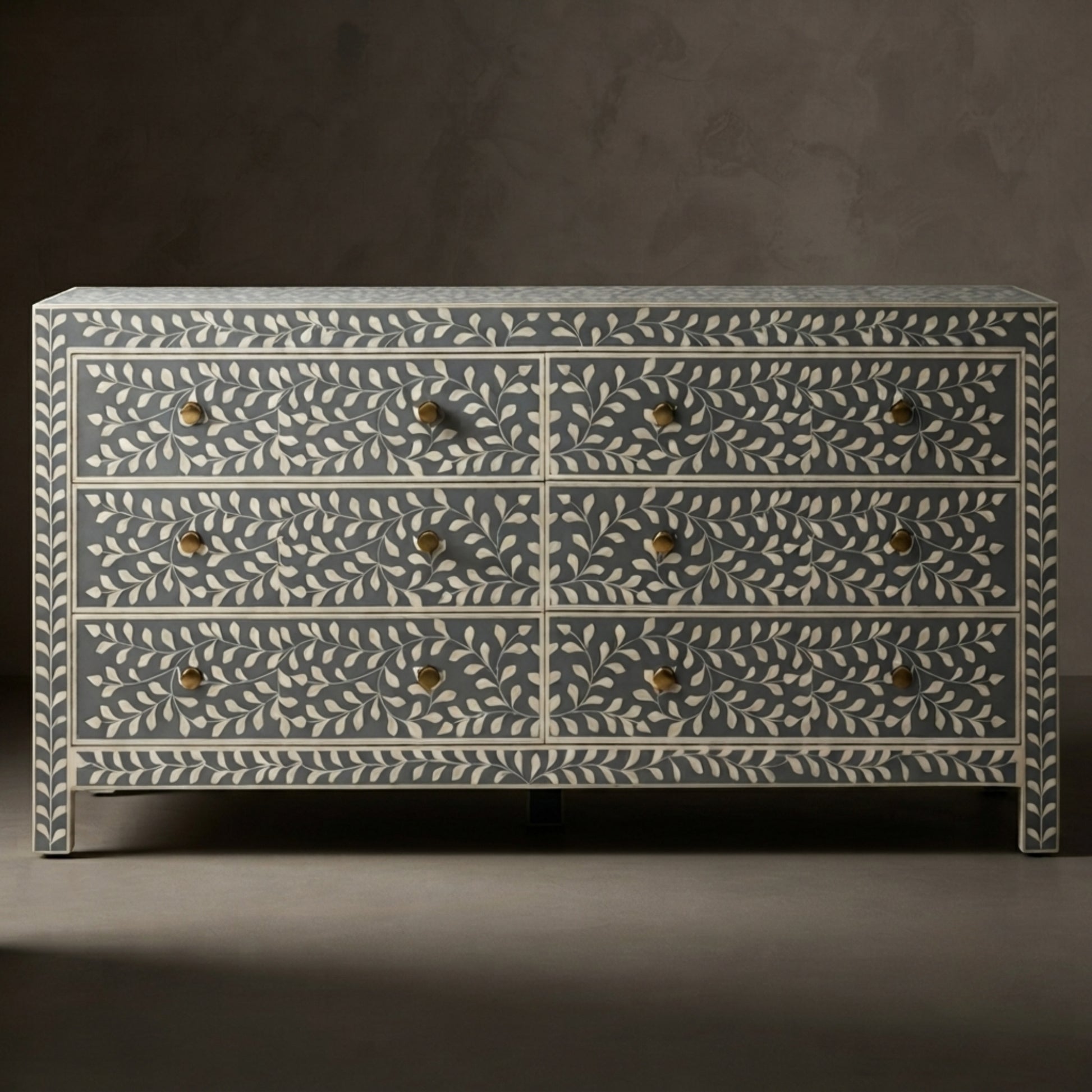 A six-drawer chest of drawers with a blue-gray and off-white bone-inlay leaf pattern and small brass handles, set against a dark gray studio background.