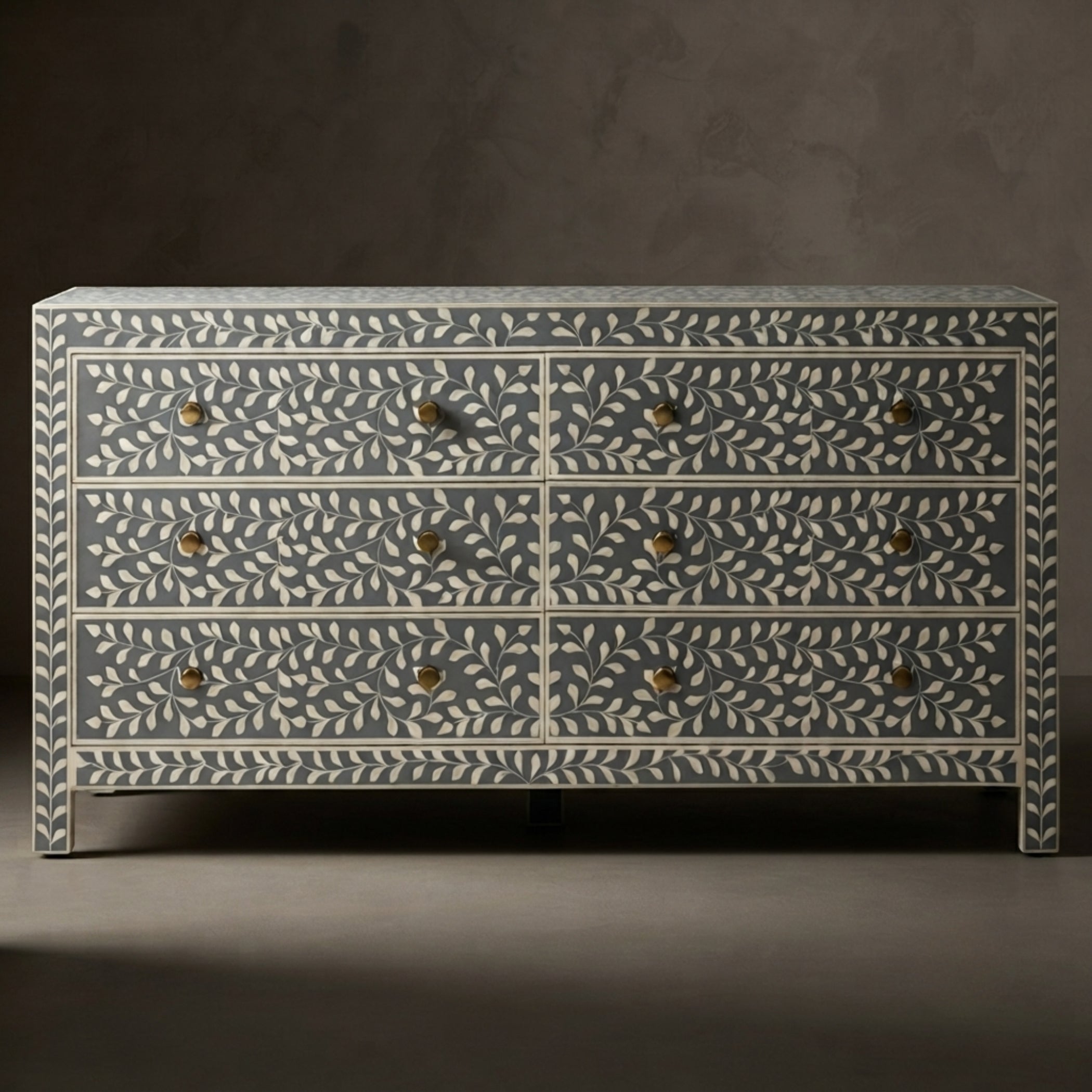 A six-drawer chest of drawers with a blue-gray and off-white bone-inlay leaf pattern and small brass handles, set against a dark gray studio background.