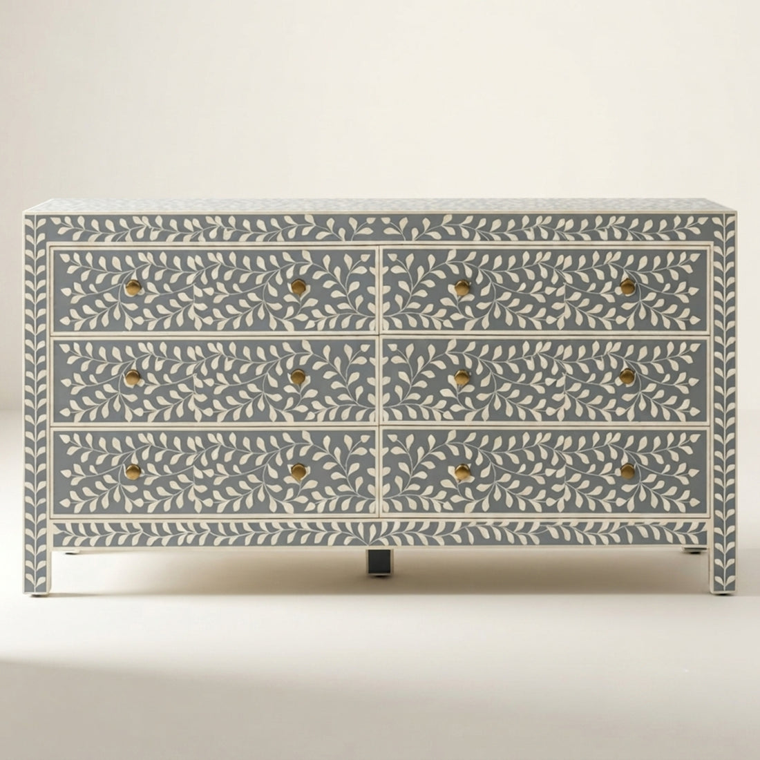 A six-drawer chest of drawers with a blue-gray and cream bone-inlay pattern and brass knobs, styled in a minimalist room setting with a wall sconce and a small vase on top.
